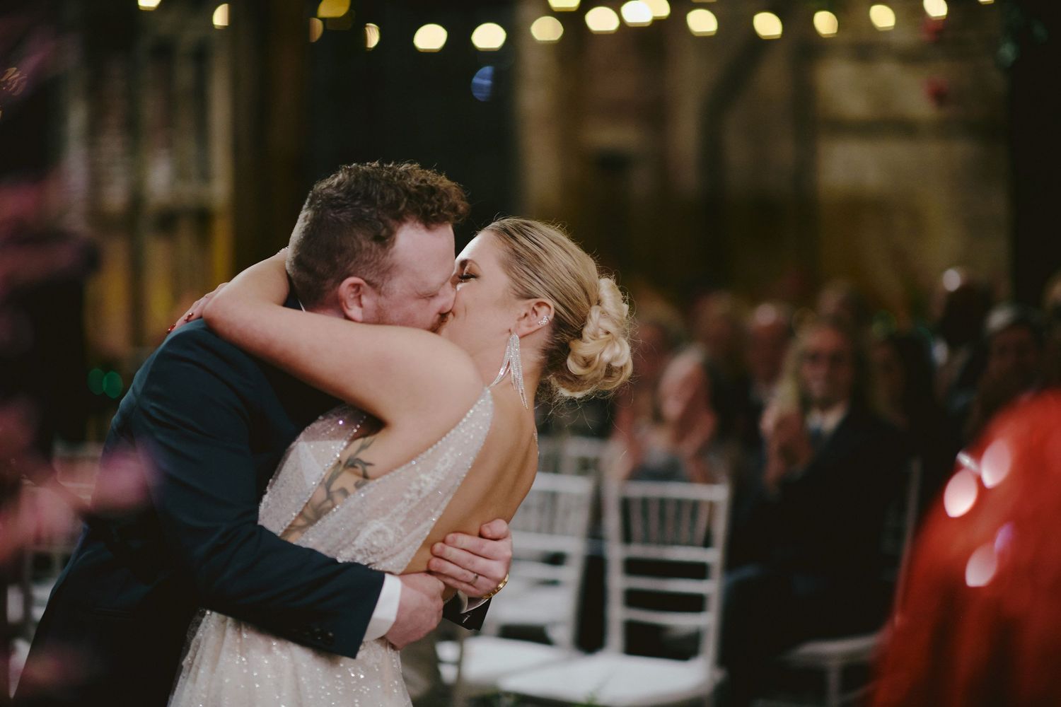 A romantic wedding couple shares an intimate kiss during their evening reception with twinkling lights in the background.