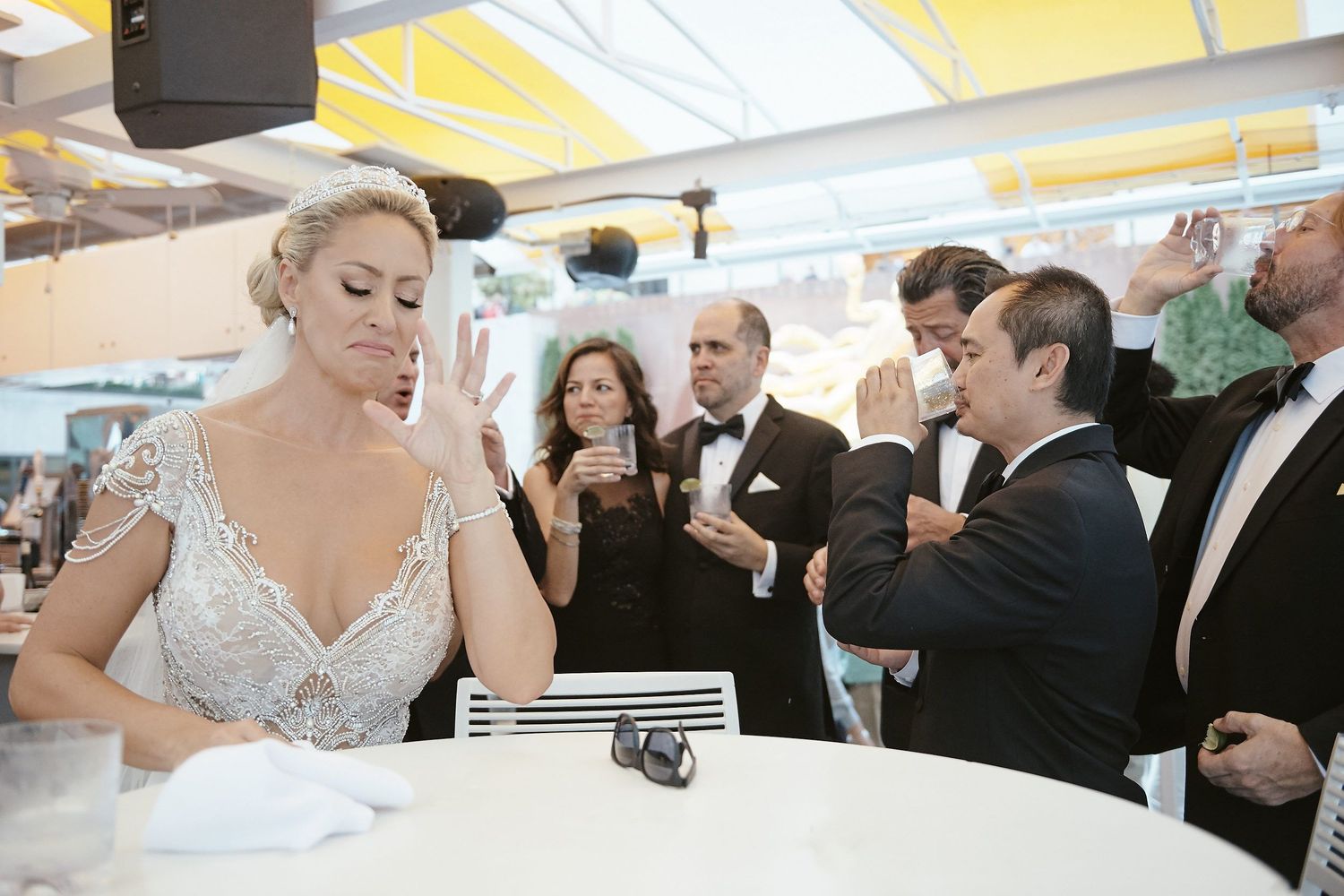 A bride in a white lace dress stands at a reception table while guests in formal attire gather around.