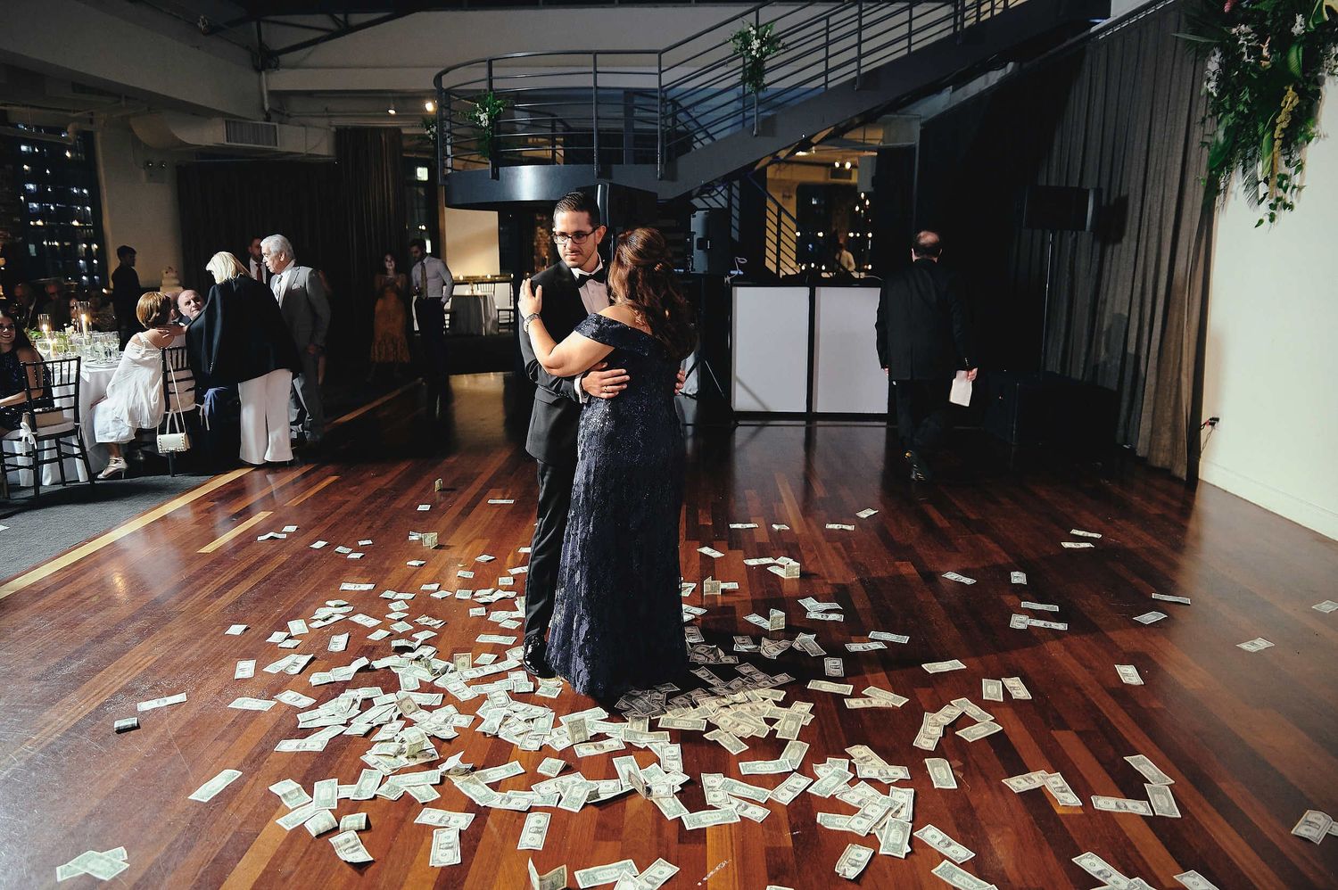 A couple slow dances together on a hardwood floor covered with scattered white rose petals in an indoor venue.