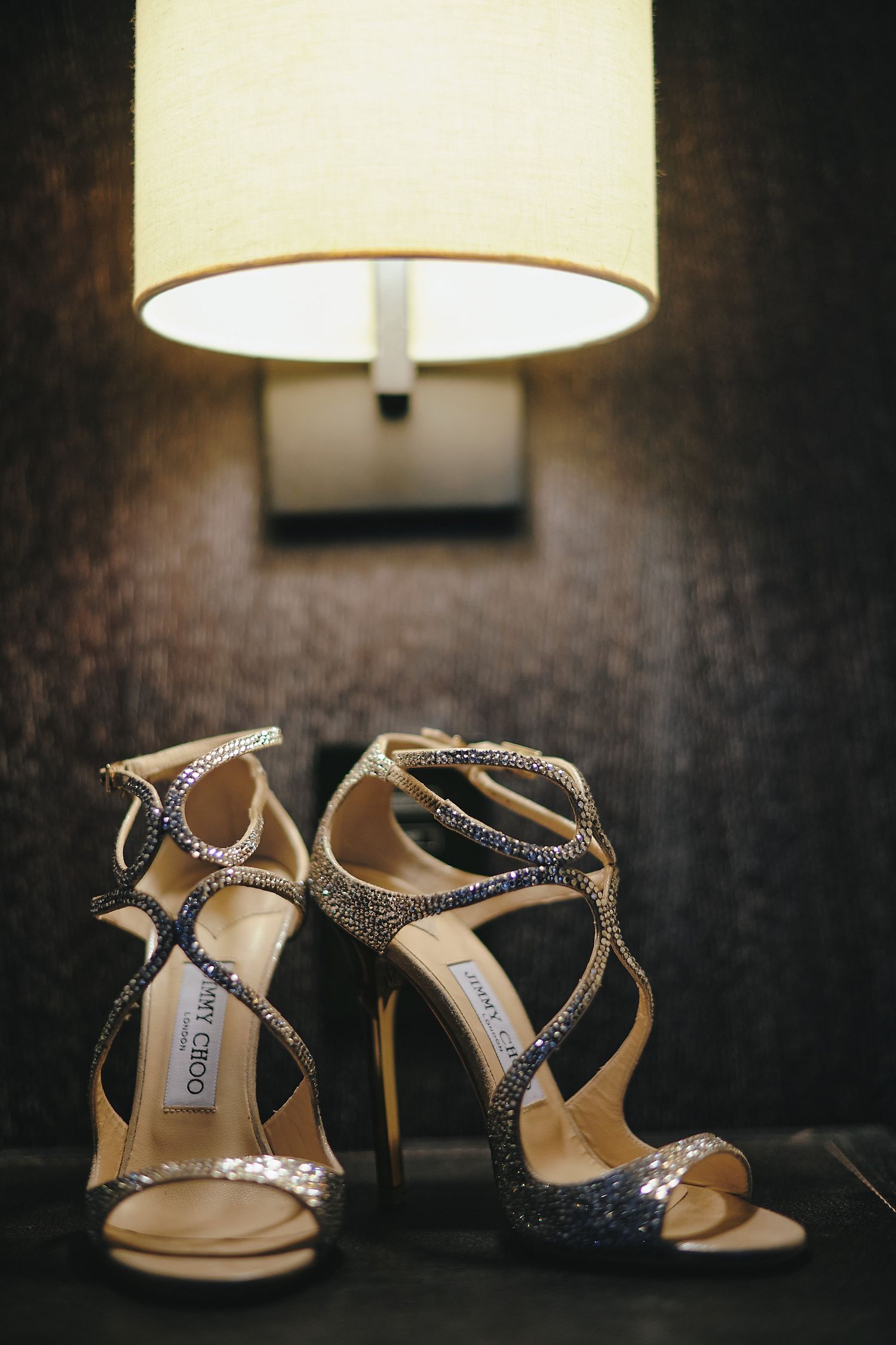 A pair of sparkly Jimmy Choo wedding sandals with crisscrossing straps sits under a warm lamp glow.