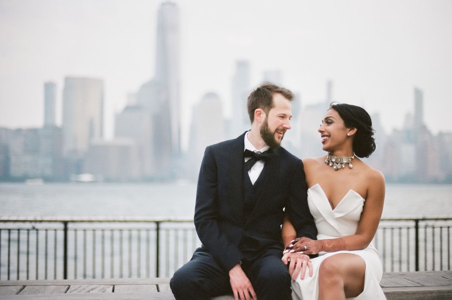 A joyful couple shares a candid moment along the waterfront with a city skyline backdrop.