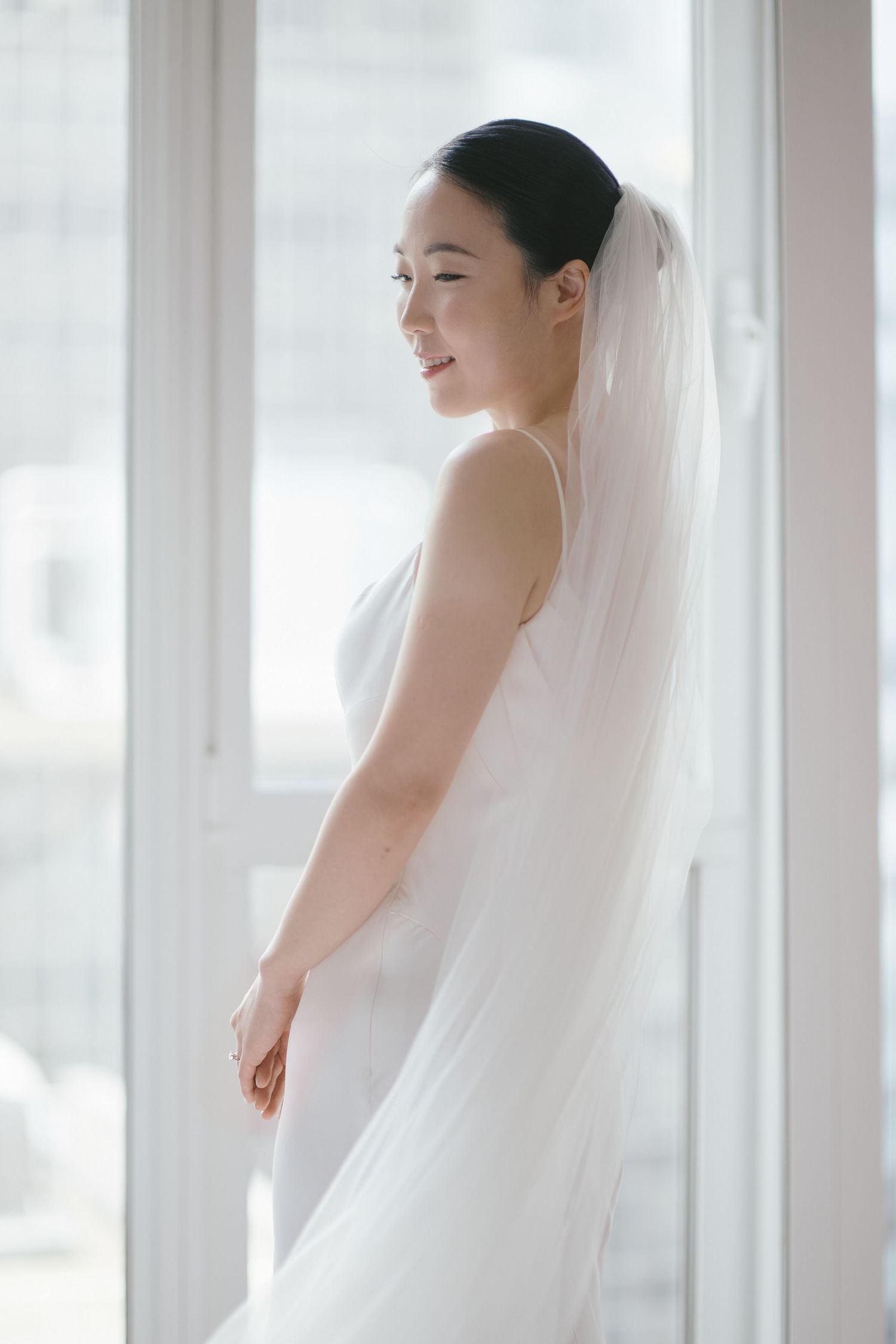 Bride in minimalist white dress with long veil poses by bright window.