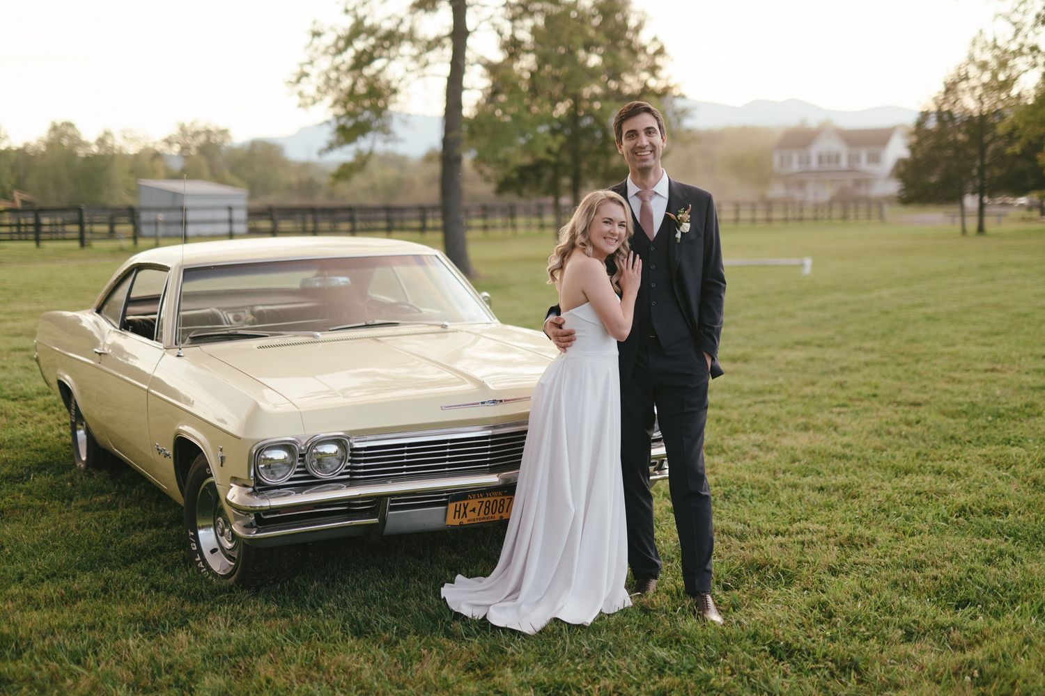 Bride and groom posing beside a vintage cream Chevrolet Impala on a green lawn at golden hour.