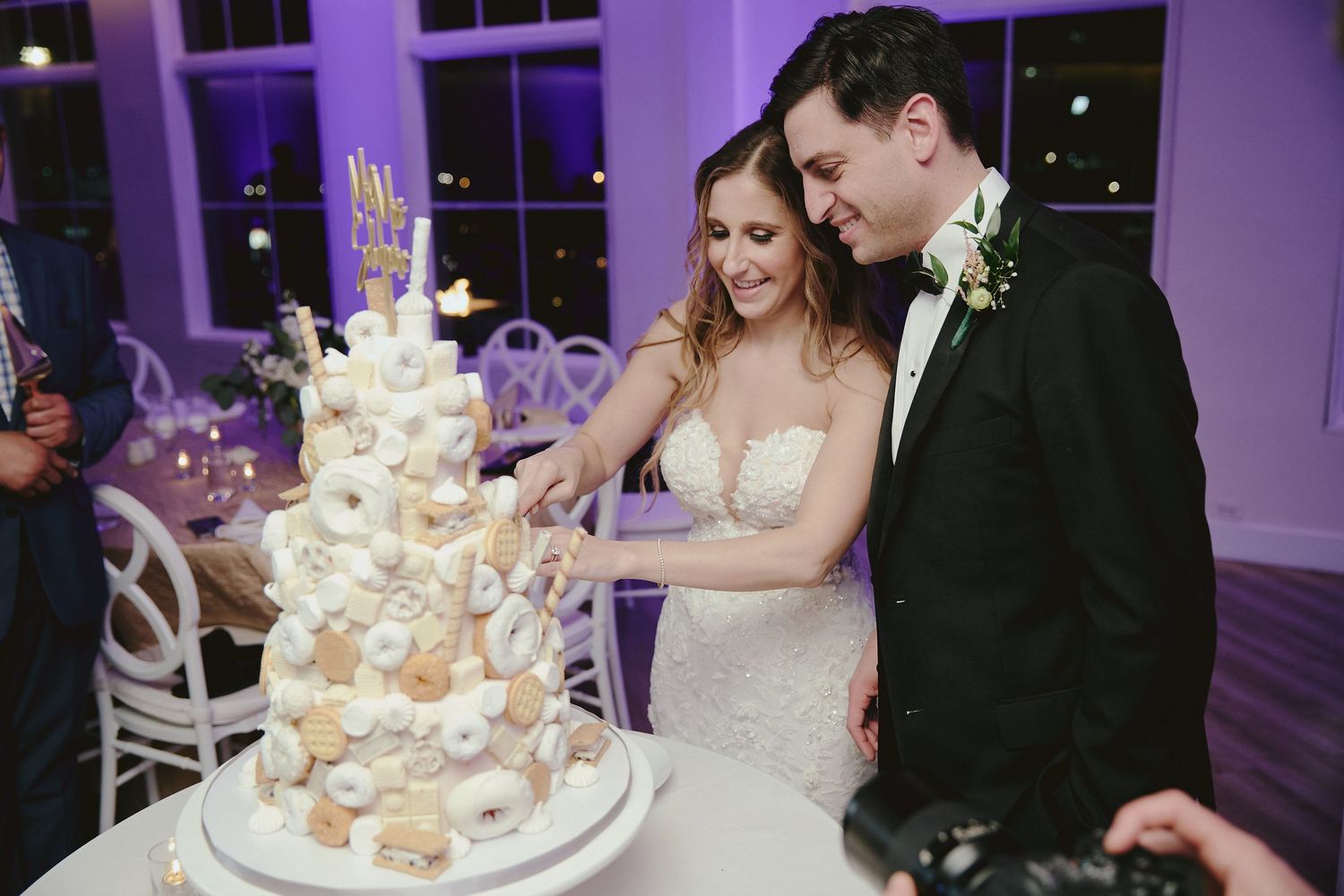 Happy couple in formal attire stands together at their wedding celebration near an elegant white tiered cake.