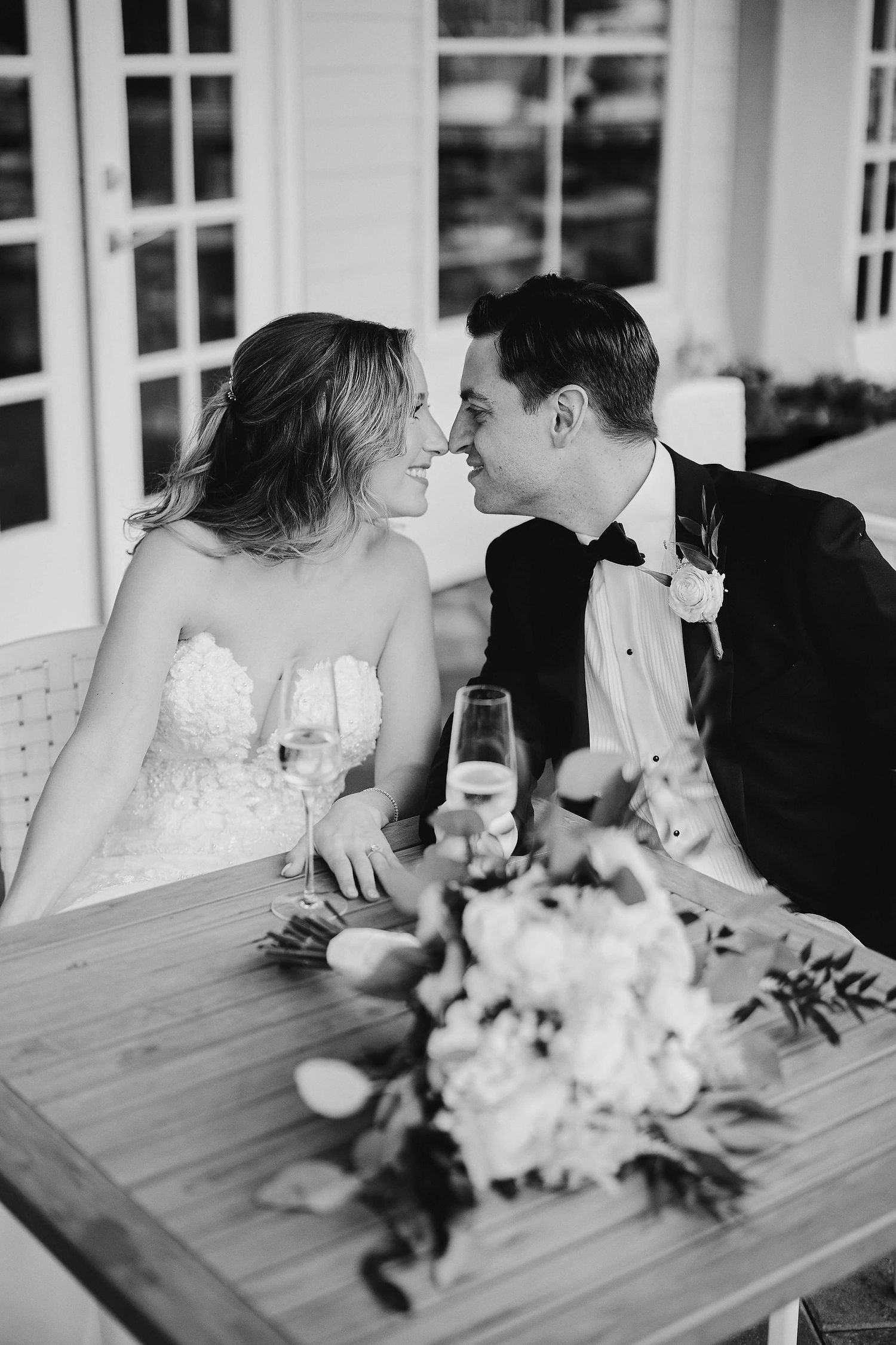 Black and white wedding photo with couple sharing intimate moment at table with champagne and floral bouquet.