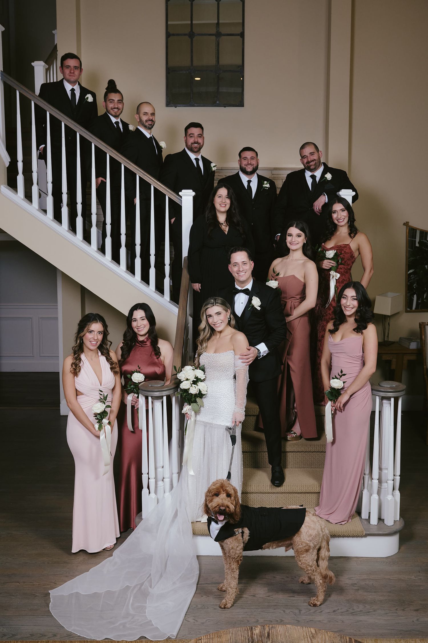 Wedding party posed on grand white staircase with groomsmen in black suits and bridesmaids in pink dresses with a dog.
