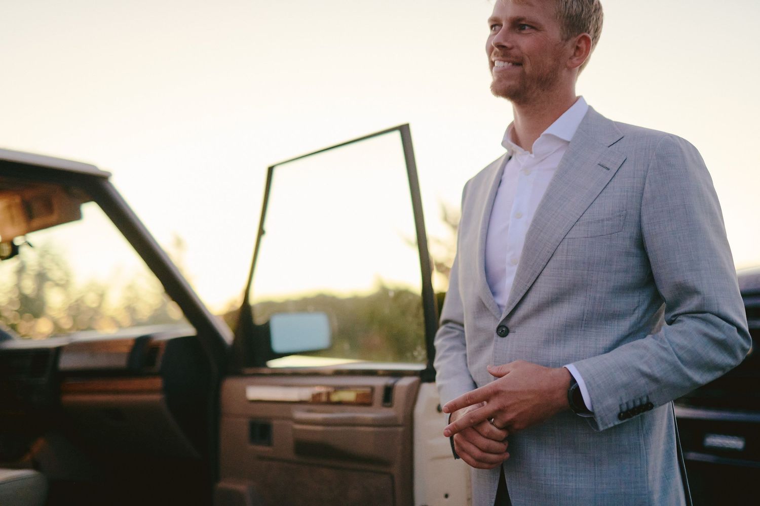 A man in a light gray suit stands beside a parked car at sunset.