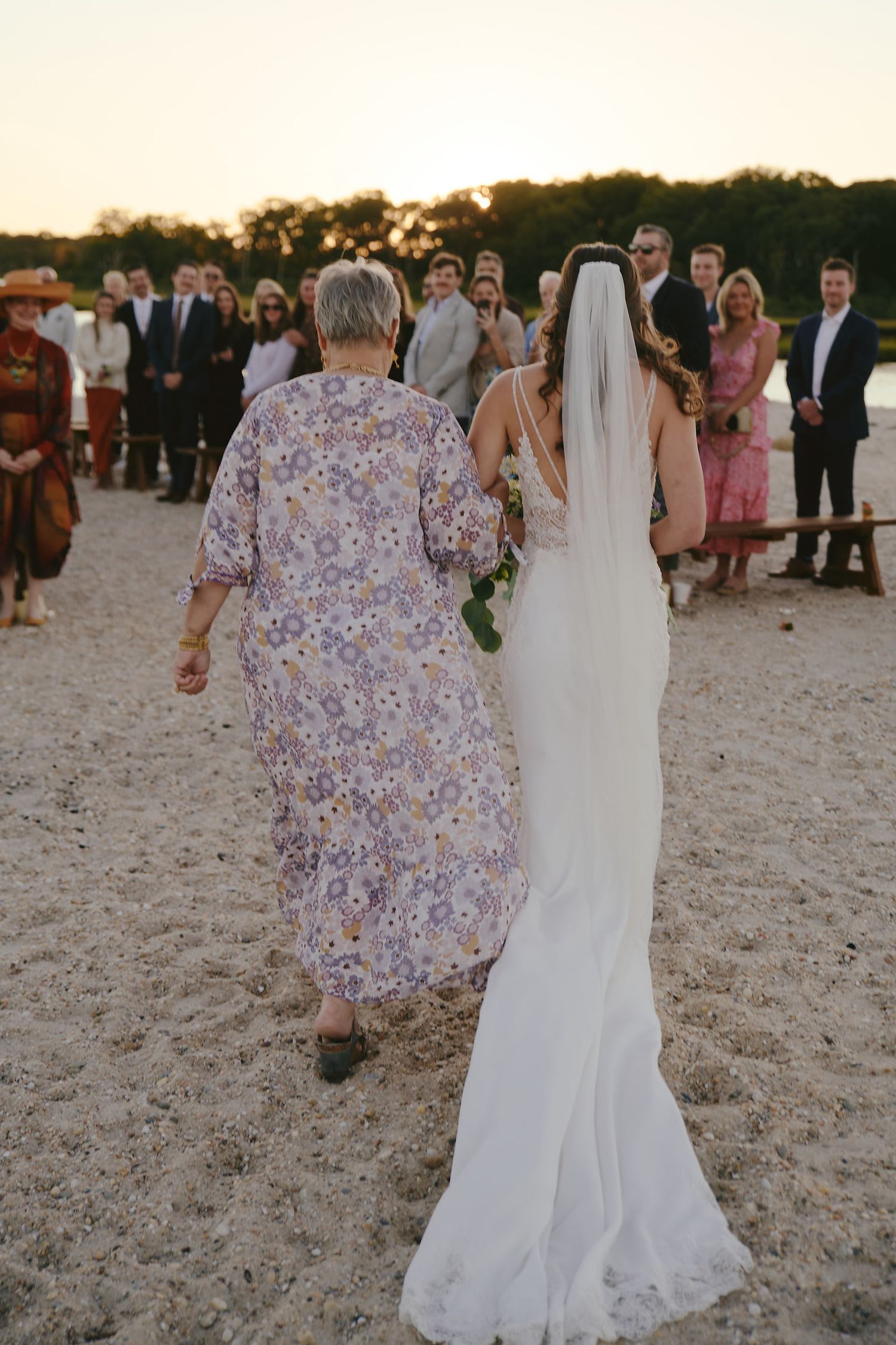 Bride in flowing white dress walks with woman in floral dress at beach wedding ceremony during sunset.