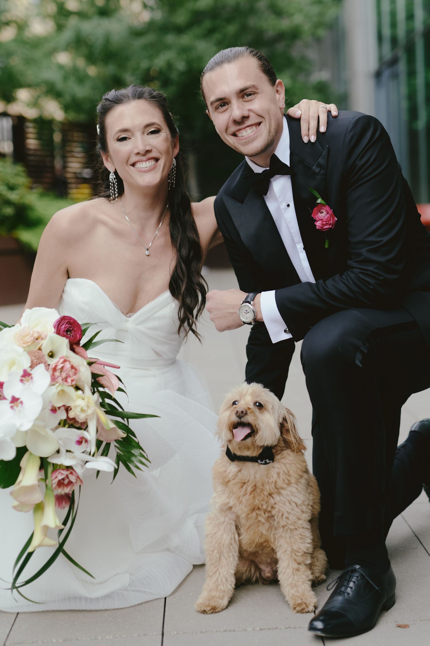 A wedding couple poses outdoors with their small fluffy dog and a beautiful white and pink floral bouquet.