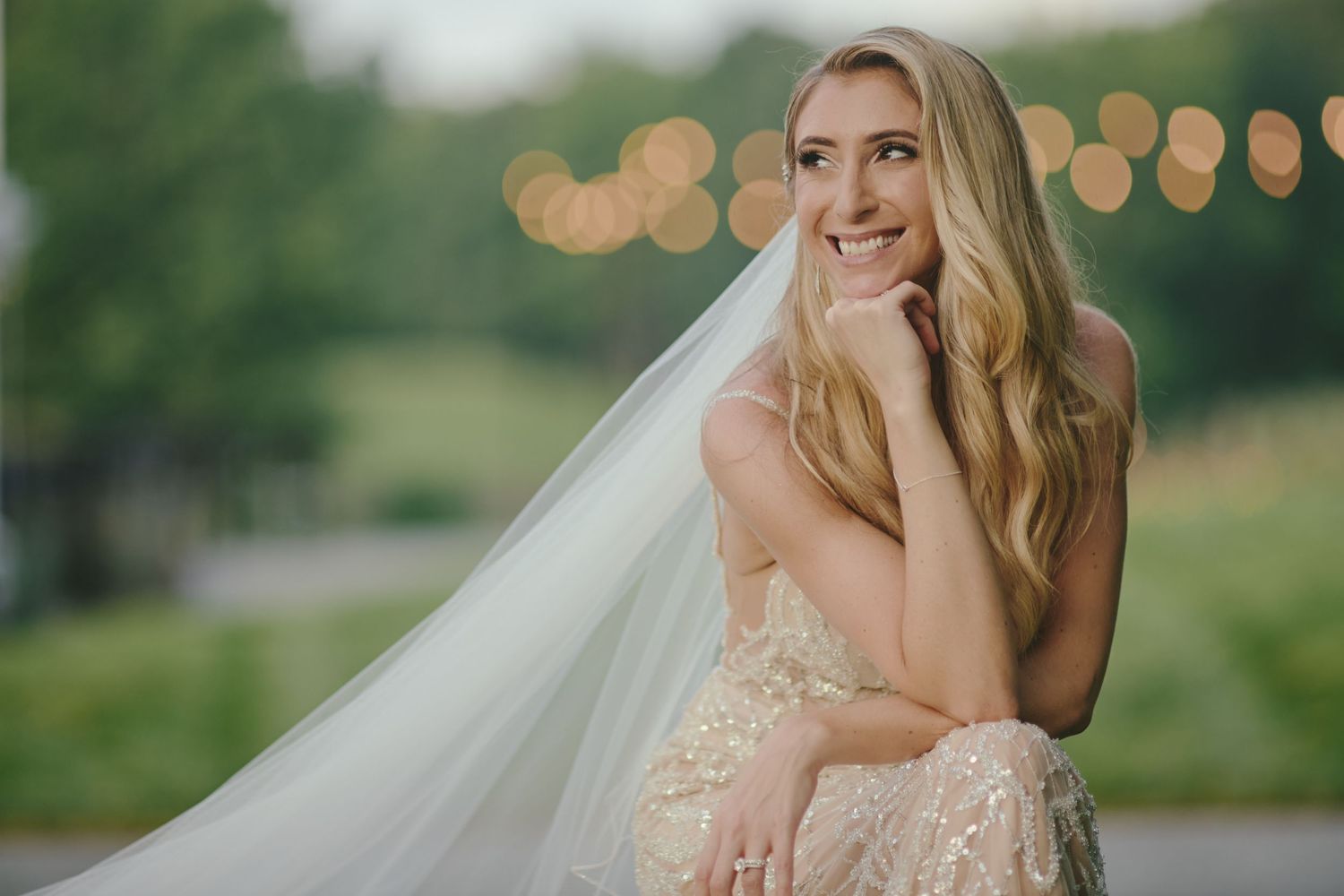 A bride in a lace wedding dress and flowing veil smiles while touching her face in an outdoor setting with bokeh lights.