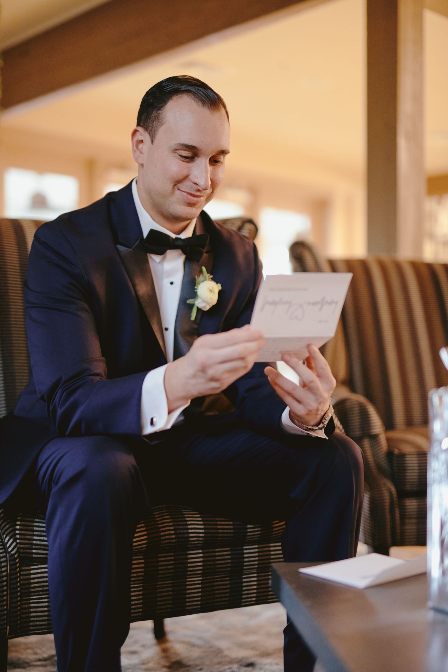 A groom in a black tuxedo and bowtie reads a letter while seated and smiling, with a white boutonniere on his lapel.