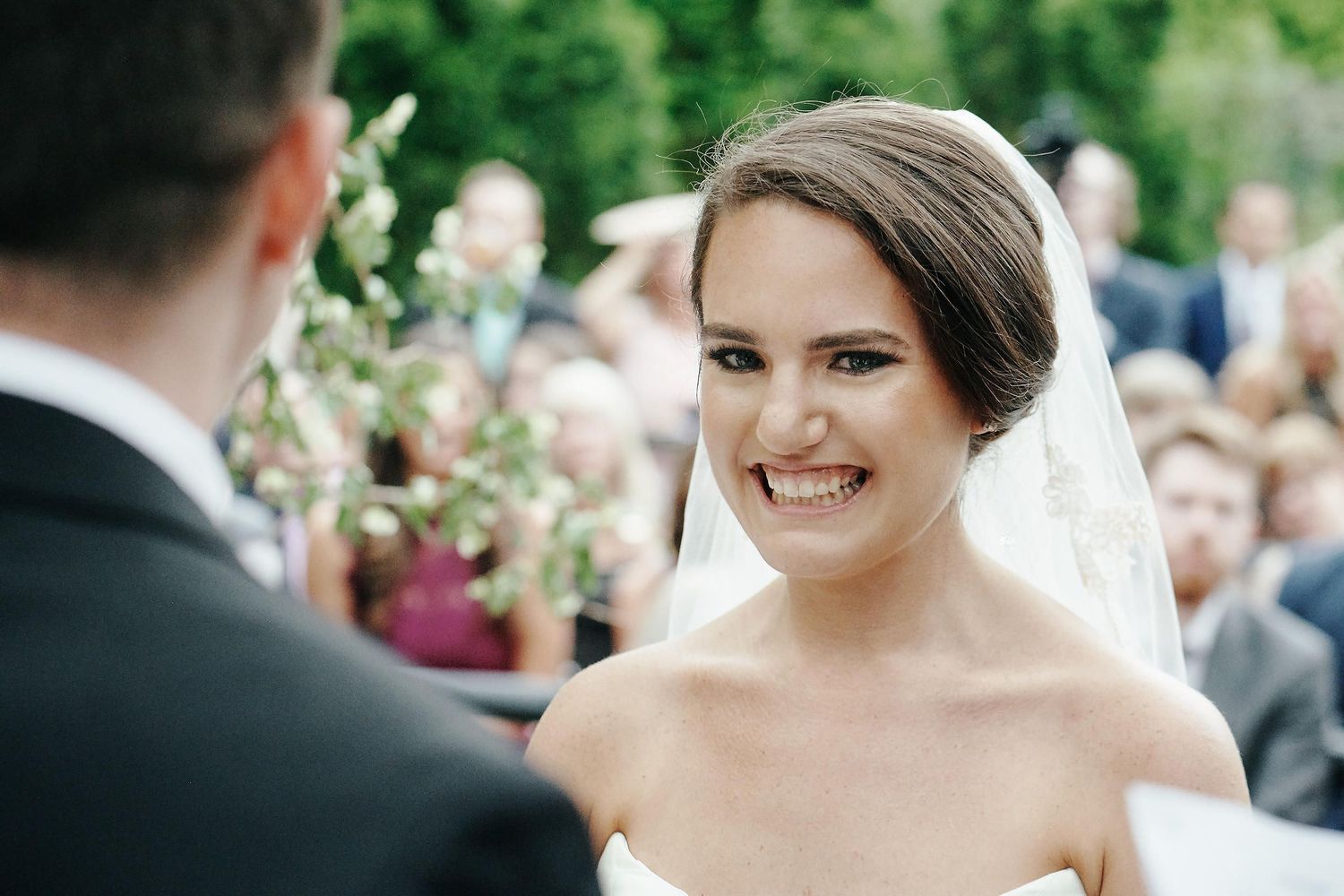A bride wearing a strapless wedding dress and veil smiling radiantly during an outdoor ceremony.