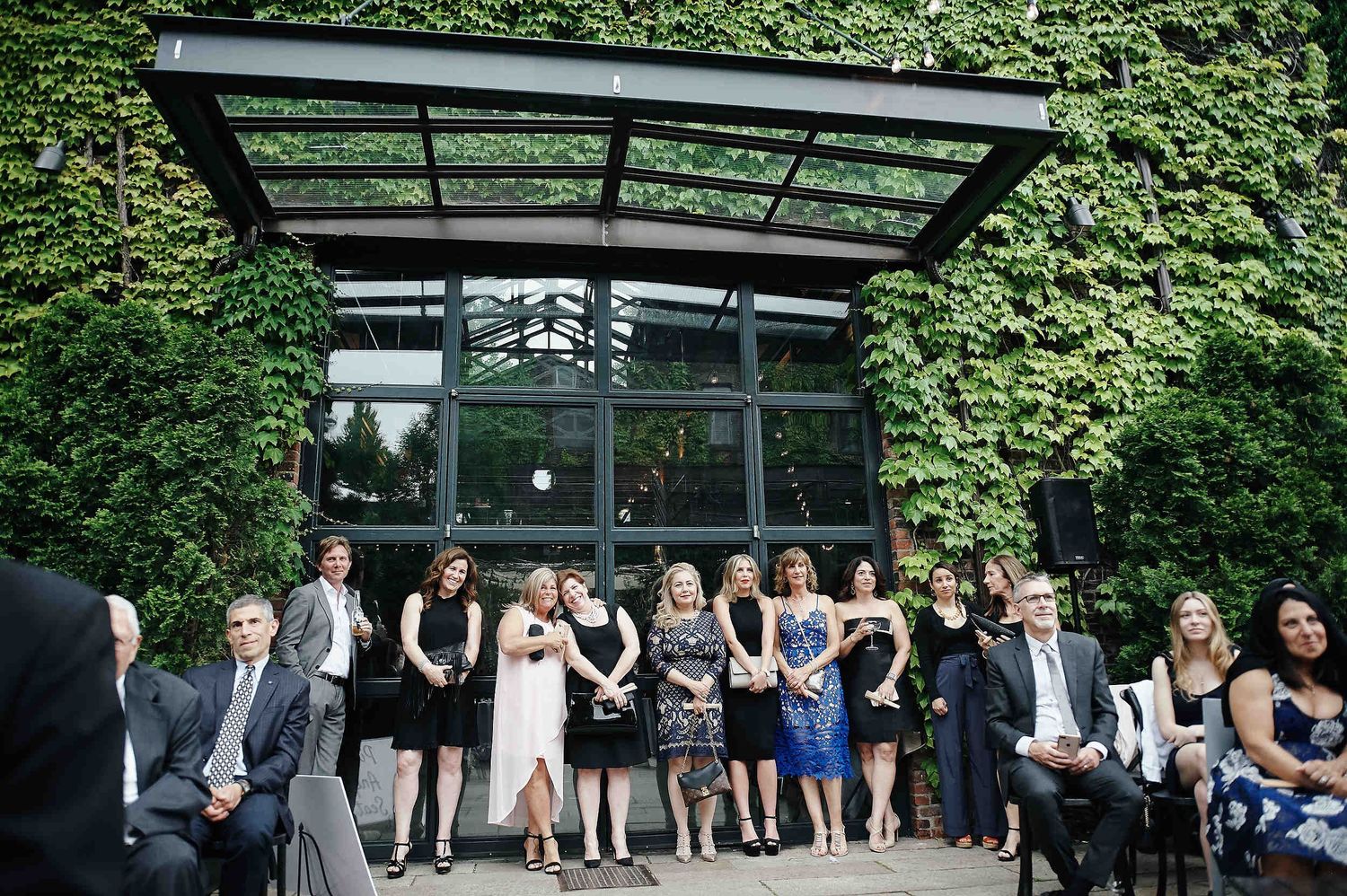 Wedding guests gather outside a modern glass building surrounded by lush green ivy-covered walls.