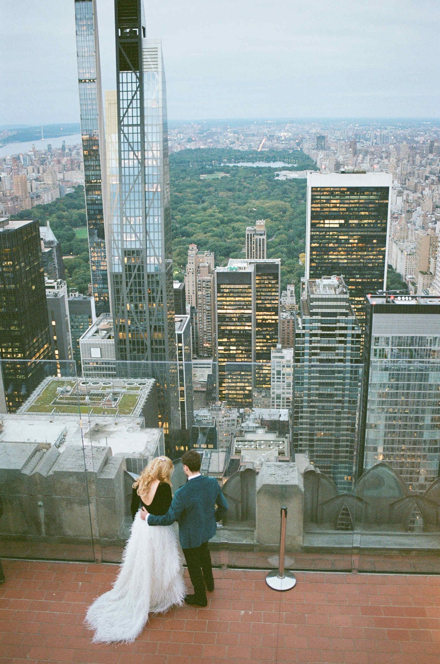 Two people admire the stunning view of Central Park and Manhattan skyline from Top of the Rock observation deck at sunset.