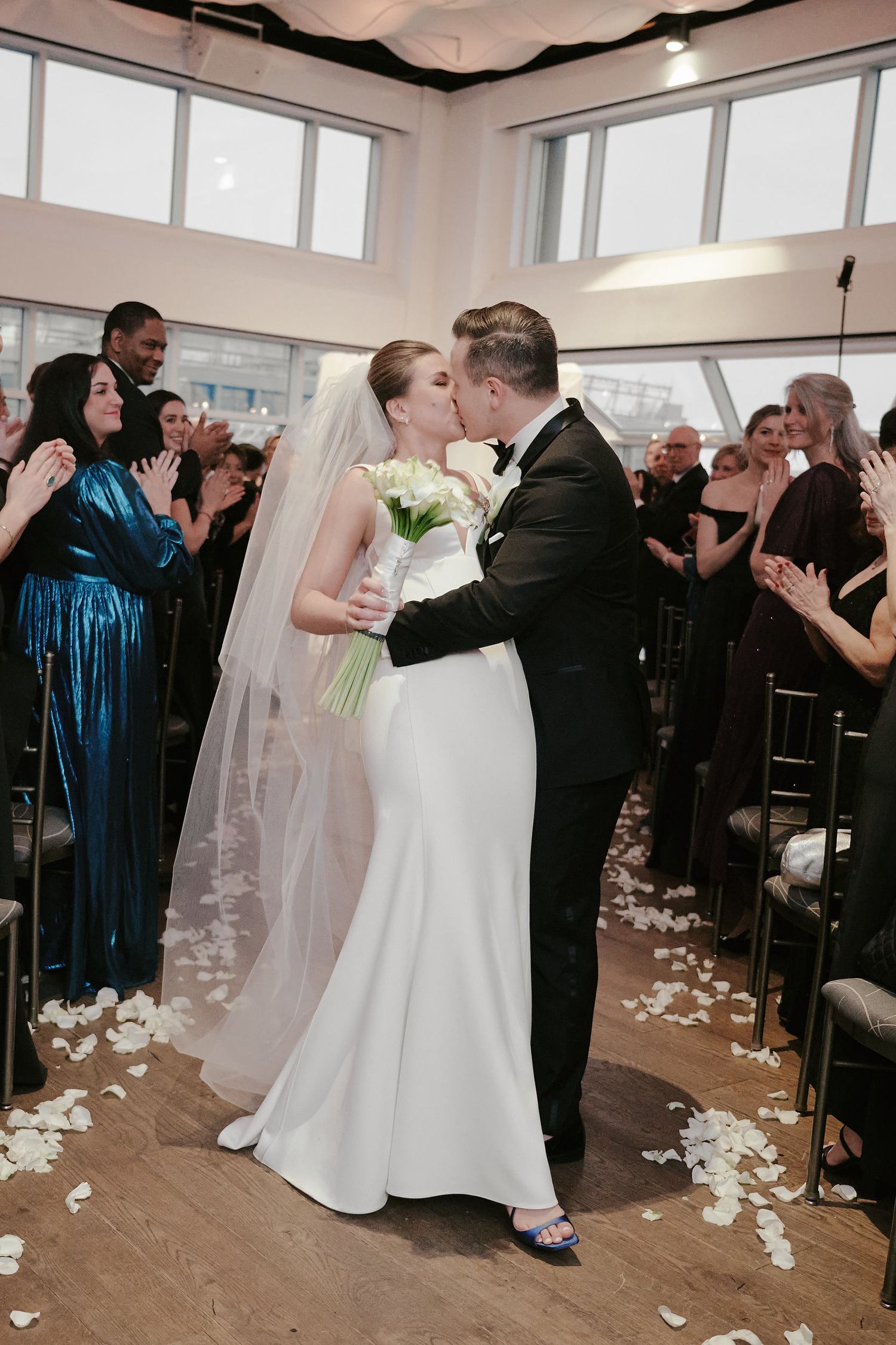 Newlyweds share their first kiss at a modern indoor ceremony as guests cheer and rose petals scatter across the hardwood floor.