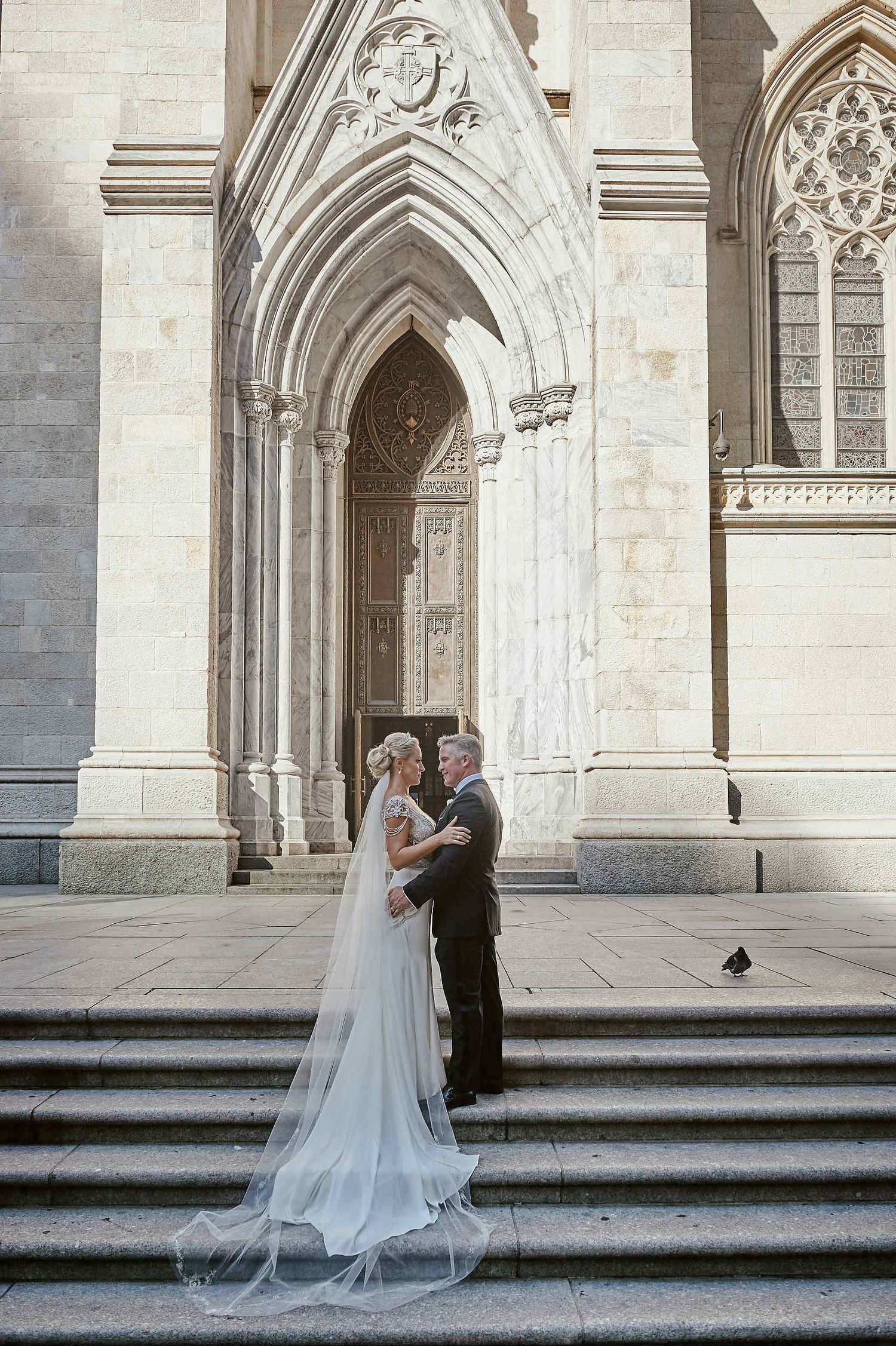 A romantic wedding couple stands on the steps of a grand Gothic cathedral with soaring arched doorways and ornate architecture.
