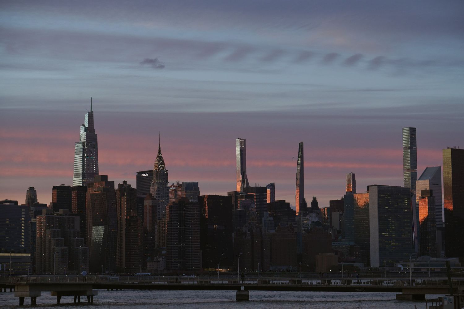 Manhattan skyline silhouetted against a dramatic purple and orange sunset sky over the East River in New York City.