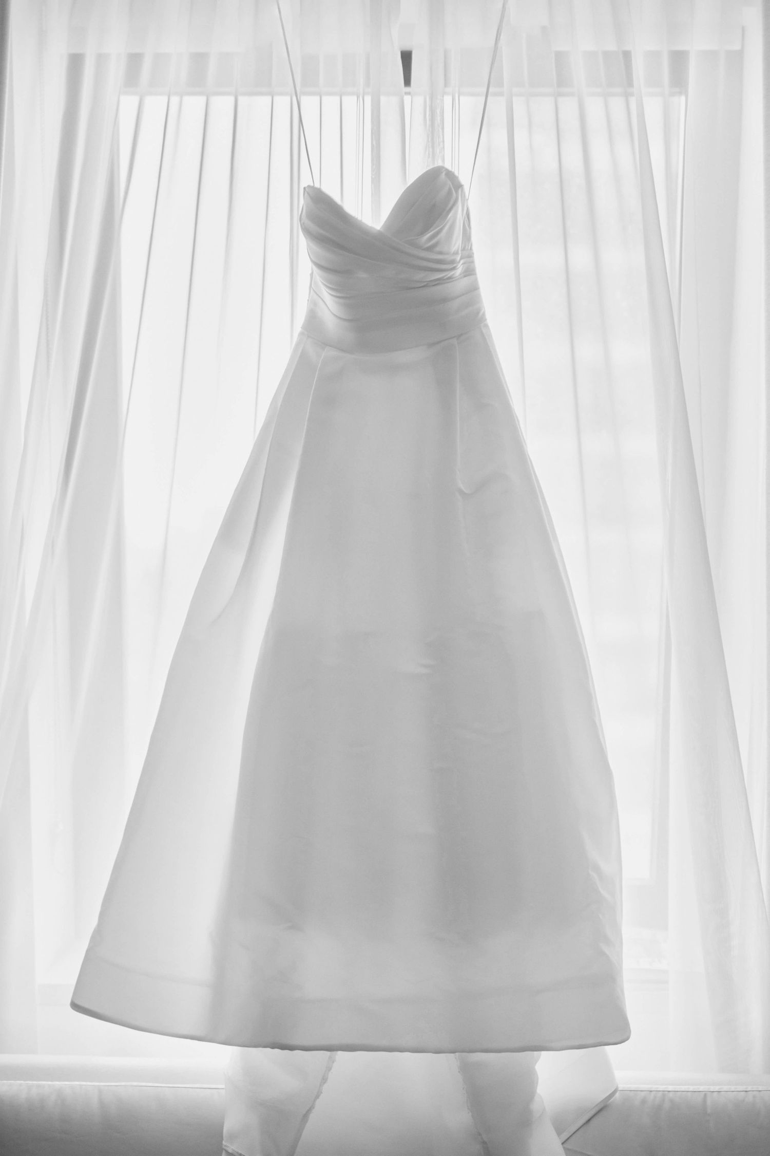 A white wedding dress hangs elegantly in front of sheer curtains, creating a soft silhouette against natural backlight.