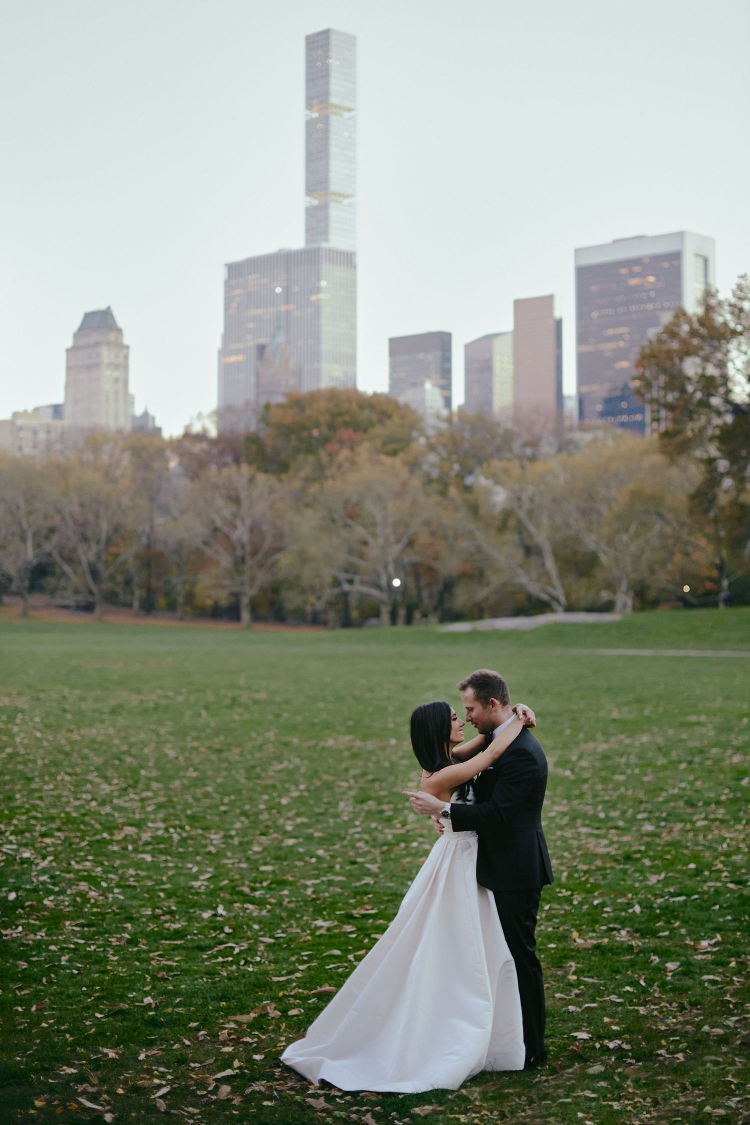 A couple in formal wedding attire share a romantic embrace on the grass in Central Park with Manhattan skyline in background.
