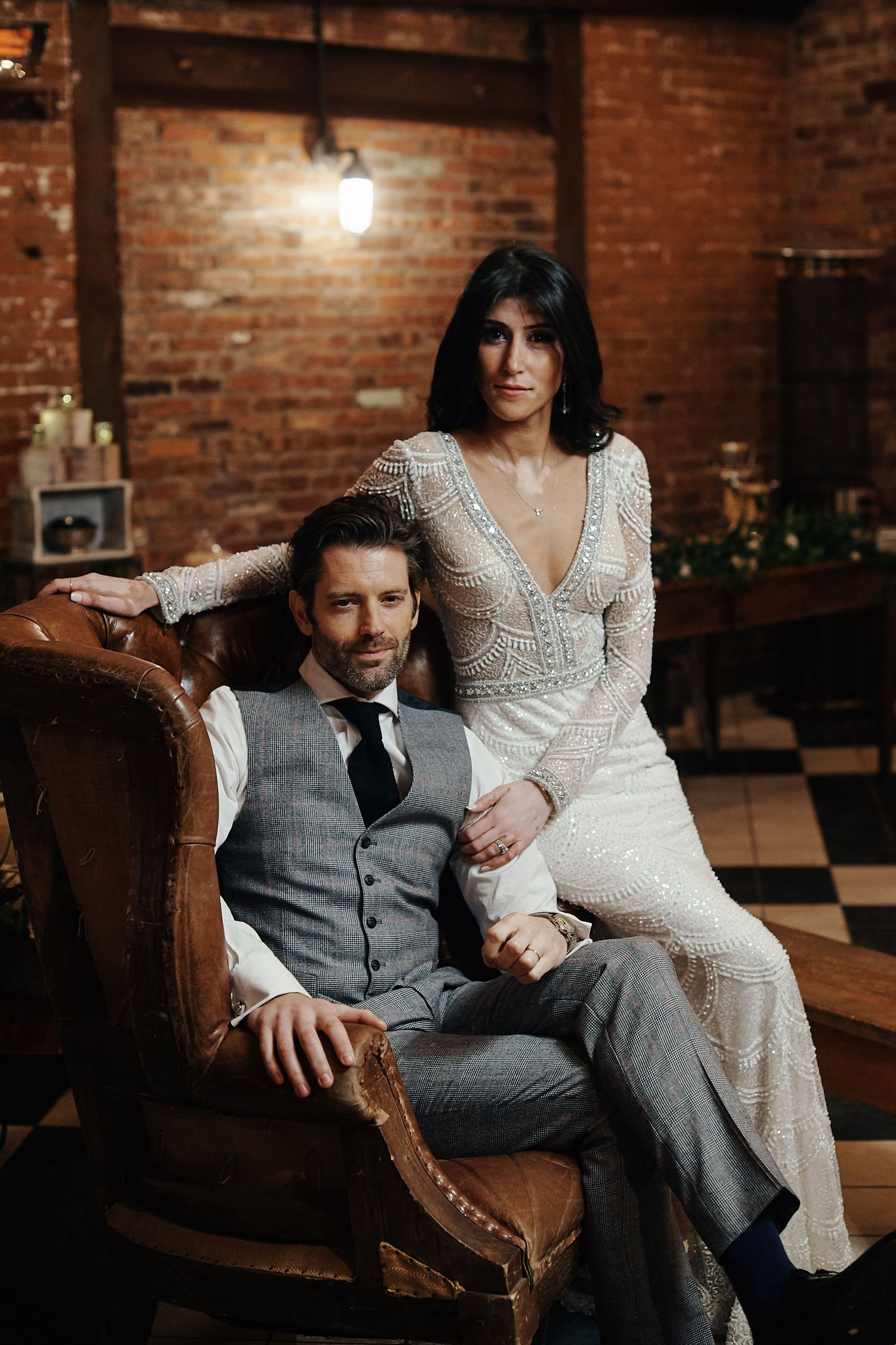 An elegant bride in a lace gown poses with a groom in a gray vest in a rustic brick venue.