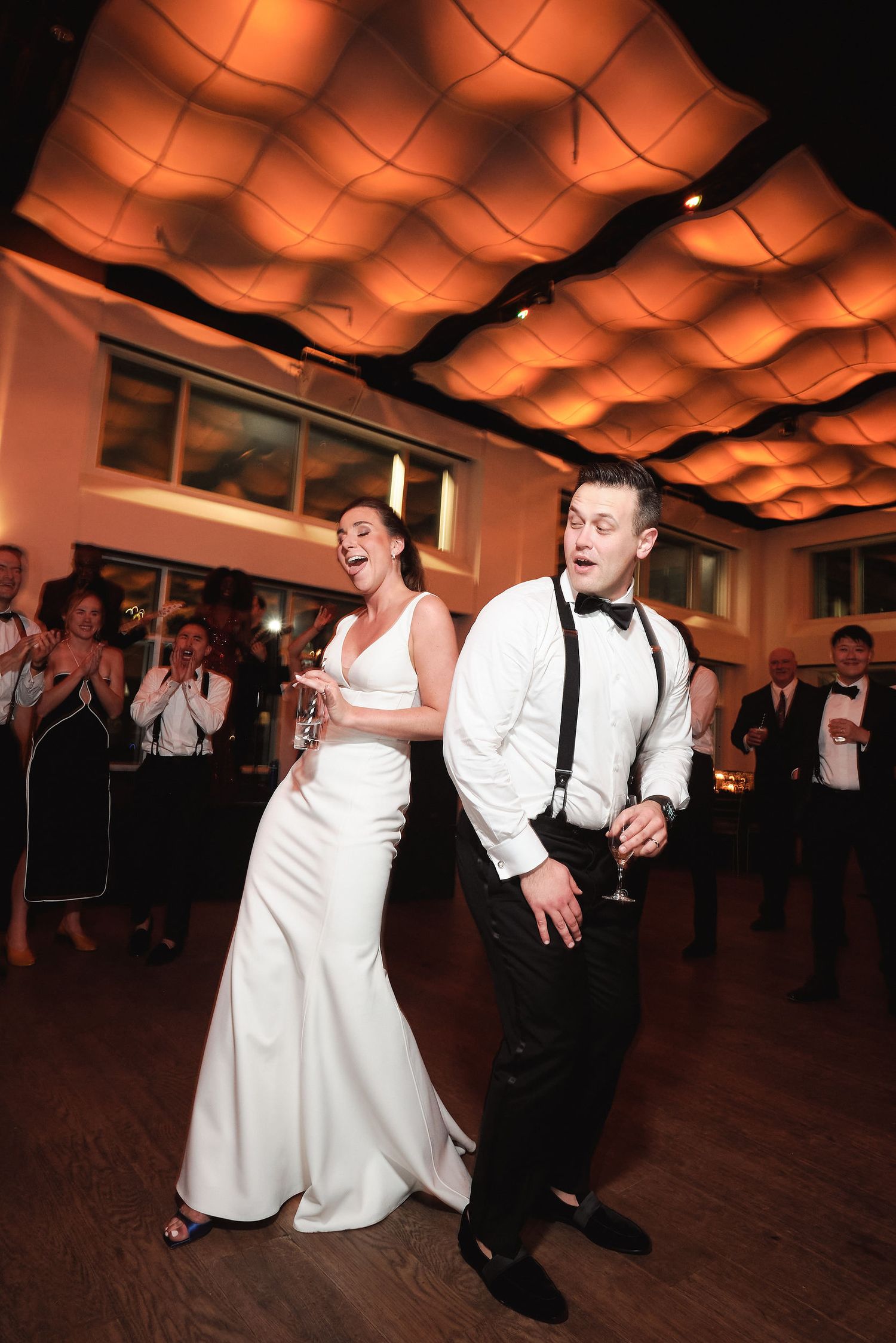A couple performs a synchronized dance move during their wedding reception under warm architectural lighting.