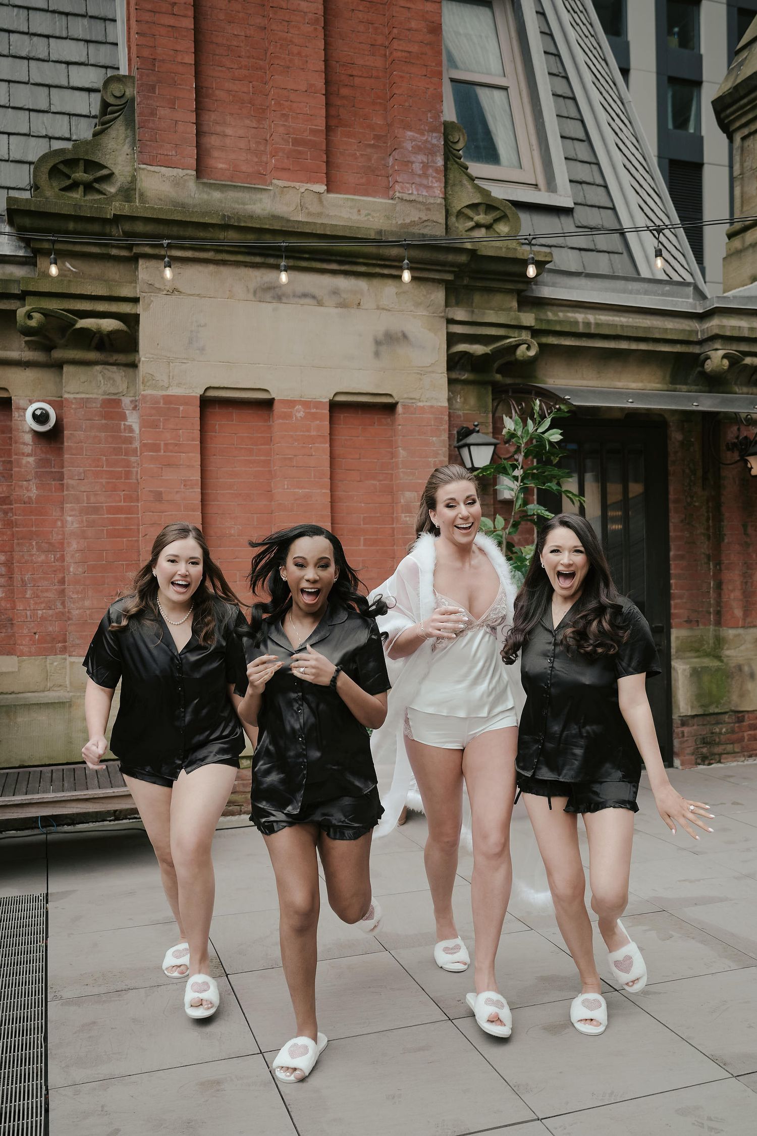 Four friends wearing black and white rompers and matching white slippers laugh and jump together outside a brick building.