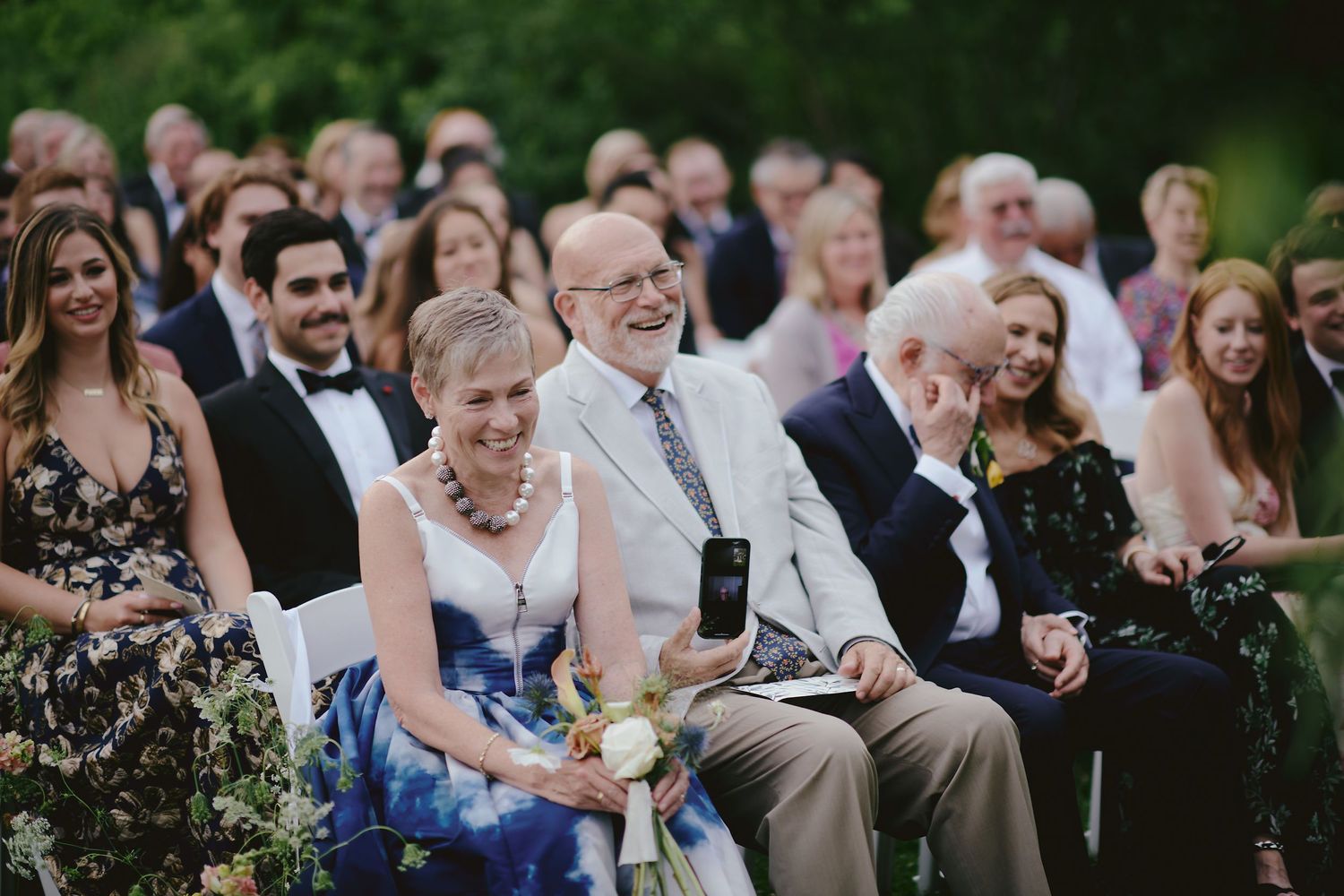 Wedding guests smile and laugh while seated in rows during an outdoor ceremony.