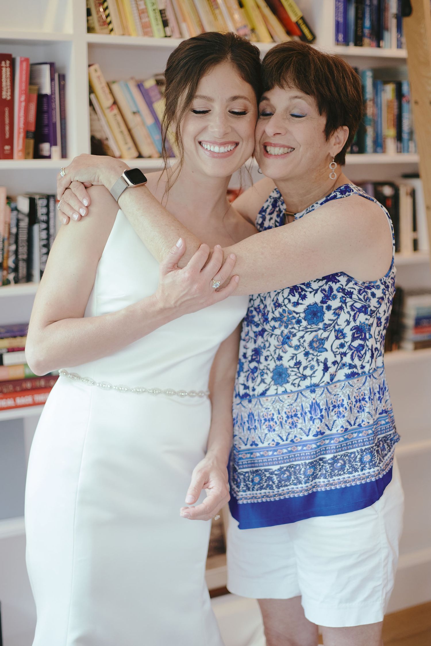 Two people share a joyful embrace in front of a bookshelf during wedding preparations.