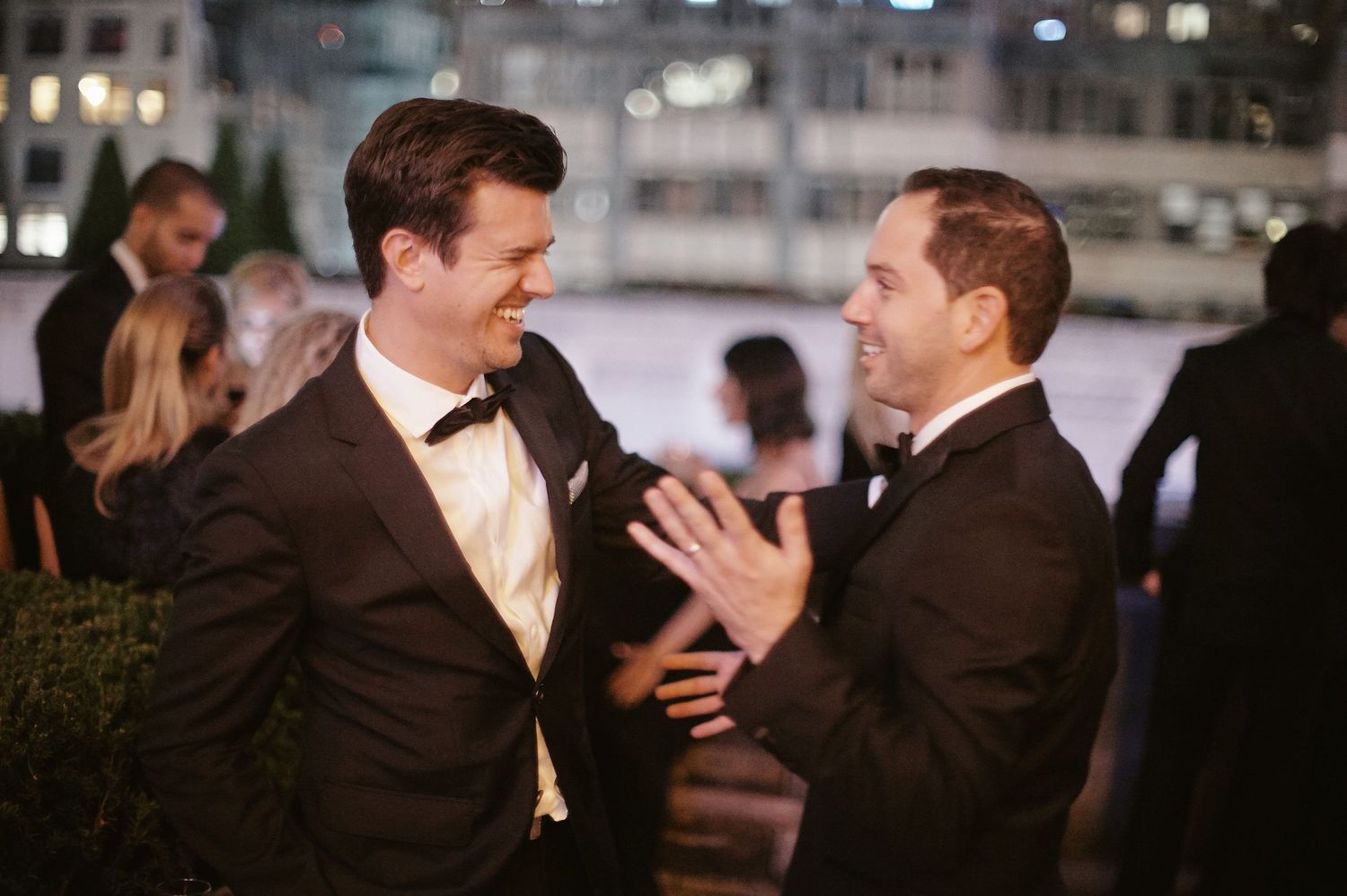 Two men in tuxedos engaged in an animated conversation at an evening event.