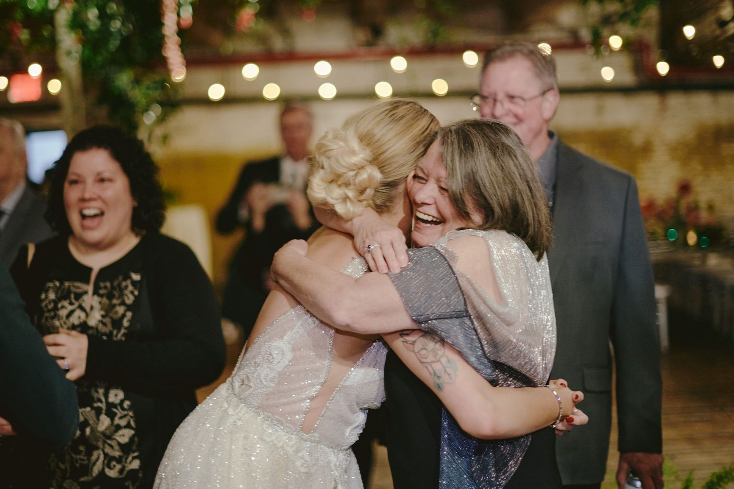 People share an emotional embrace on a dance floor at a wedding reception with string lights and greenery decorating the venue.