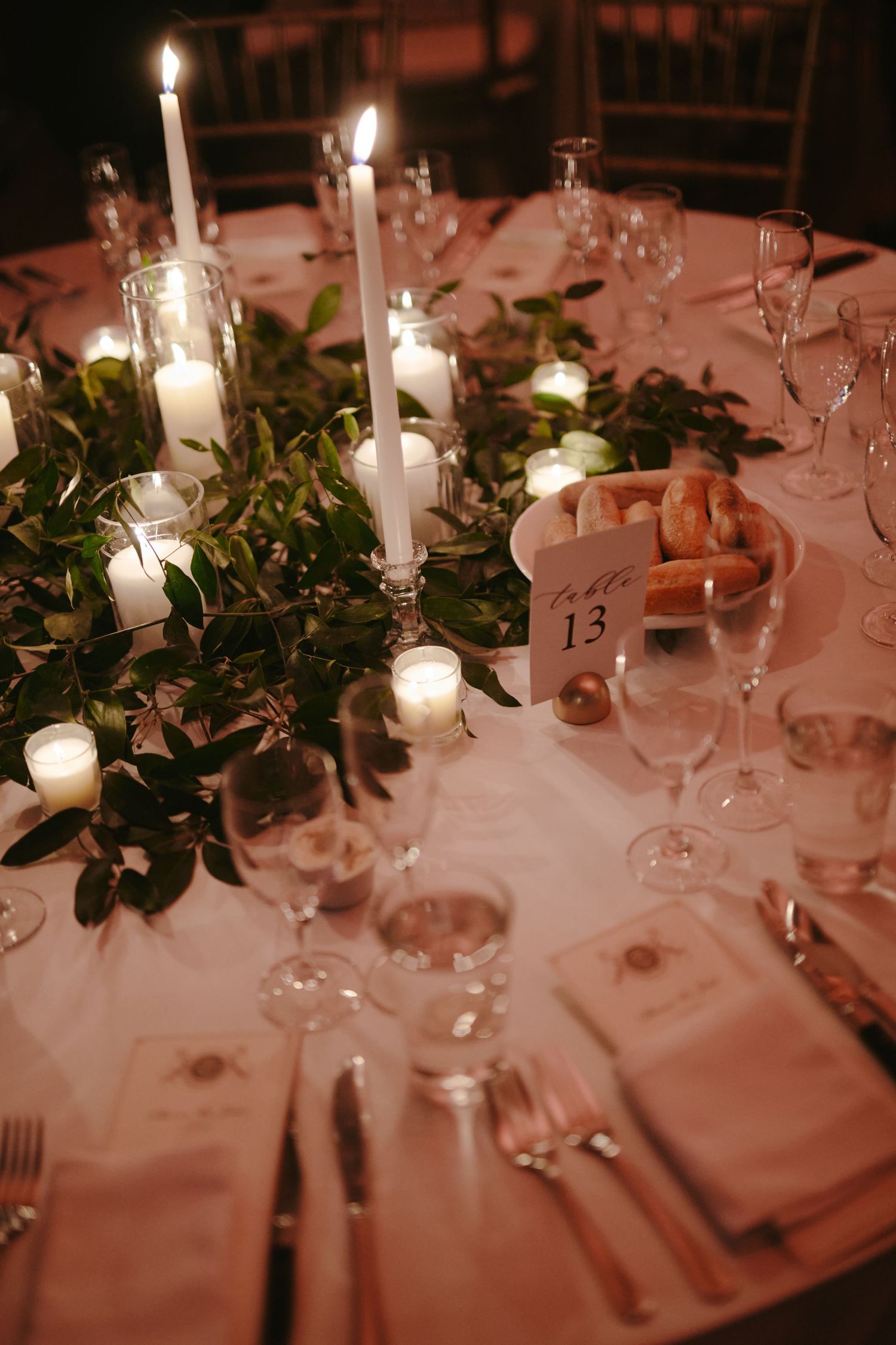 Elegant wedding table setting with white candles, greenery centerpiece, wine glasses and table number 13.