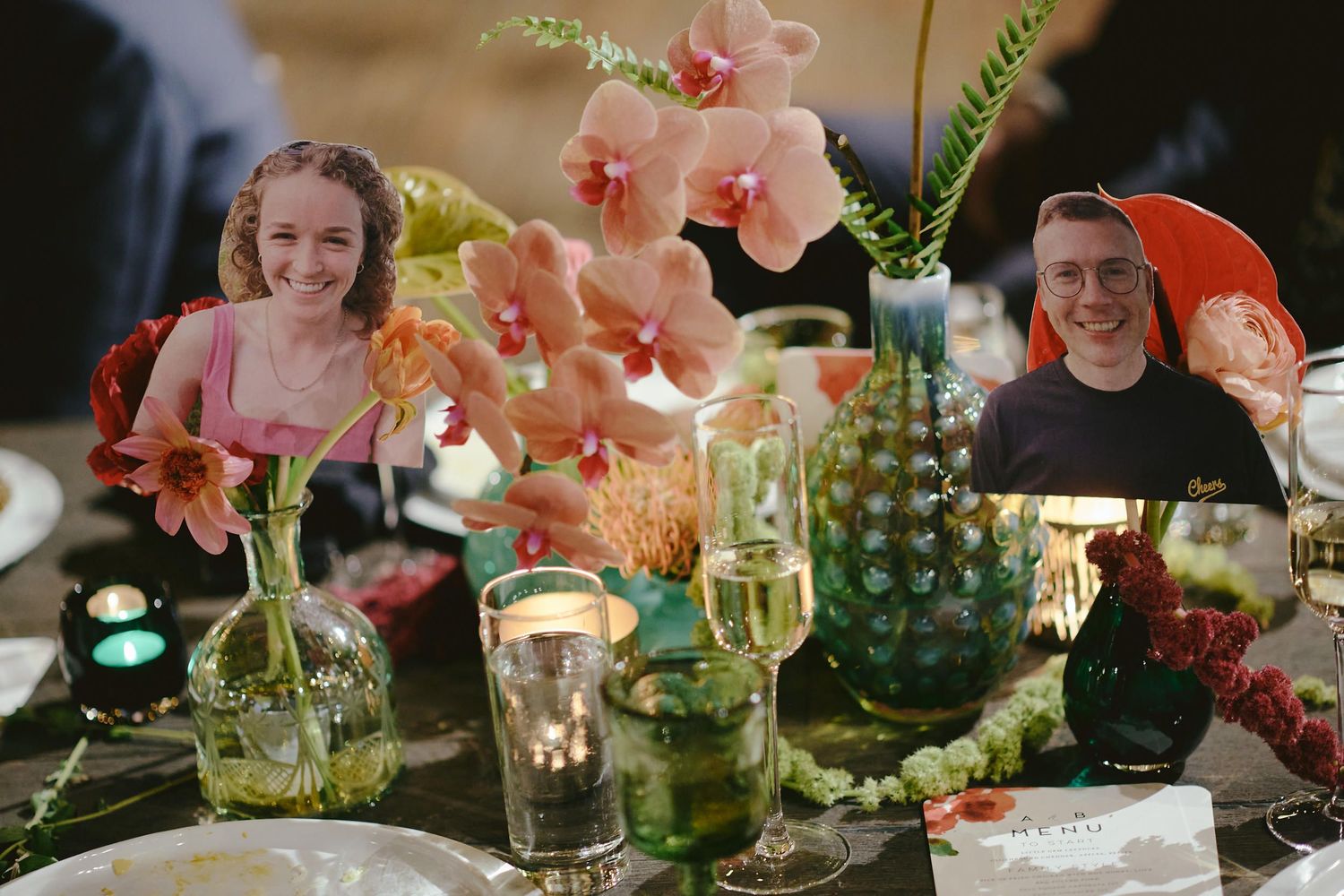 Elegant dinner table setting with pink orchids in glass vases, green glassware, and mood lighting at evening gathering.
