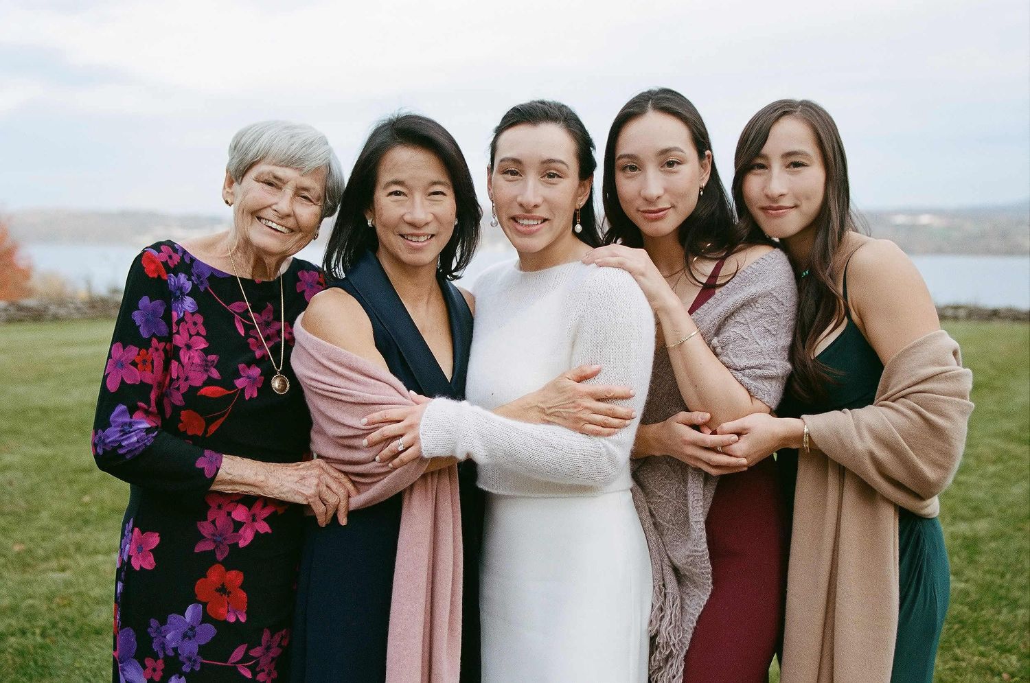 Five women dressed in elegant casual wear stand together embracing outdoors with water and cloudy sky in background.