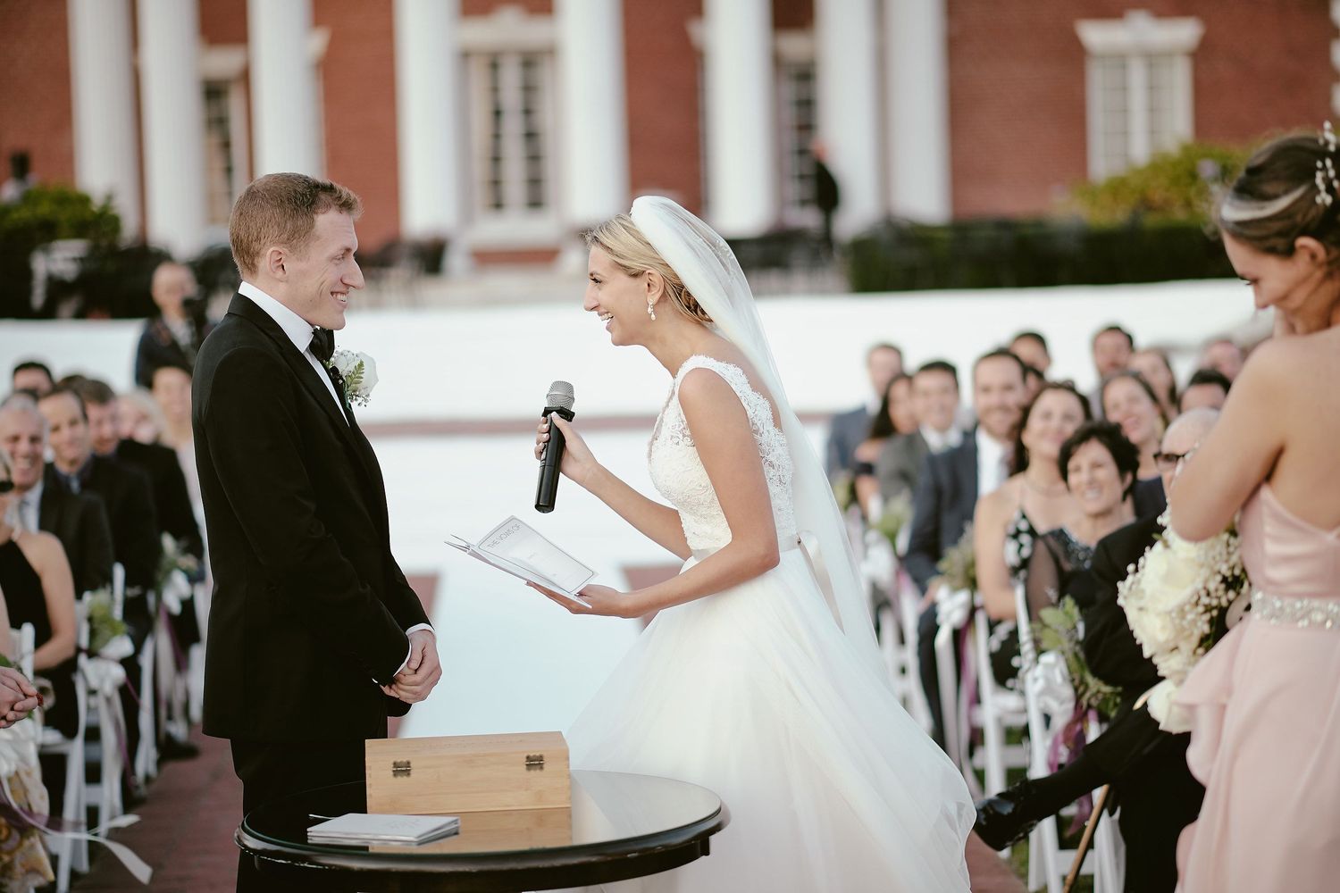 Outdoor wedding ceremony with couple exchanging vows in front of gathered guests on white chairs.