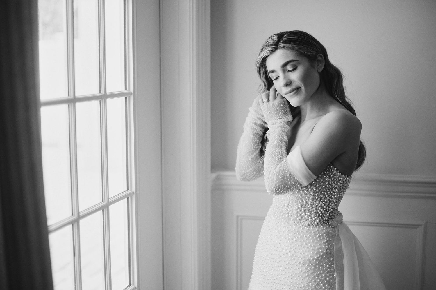 A bride in an elegant beaded wedding dress poses gracefully by a bright window in black and white.