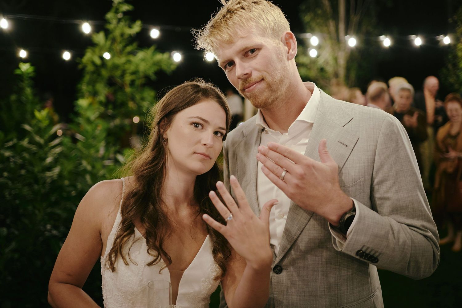 A couple shows off their wedding rings under string lights at an outdoor evening celebration.