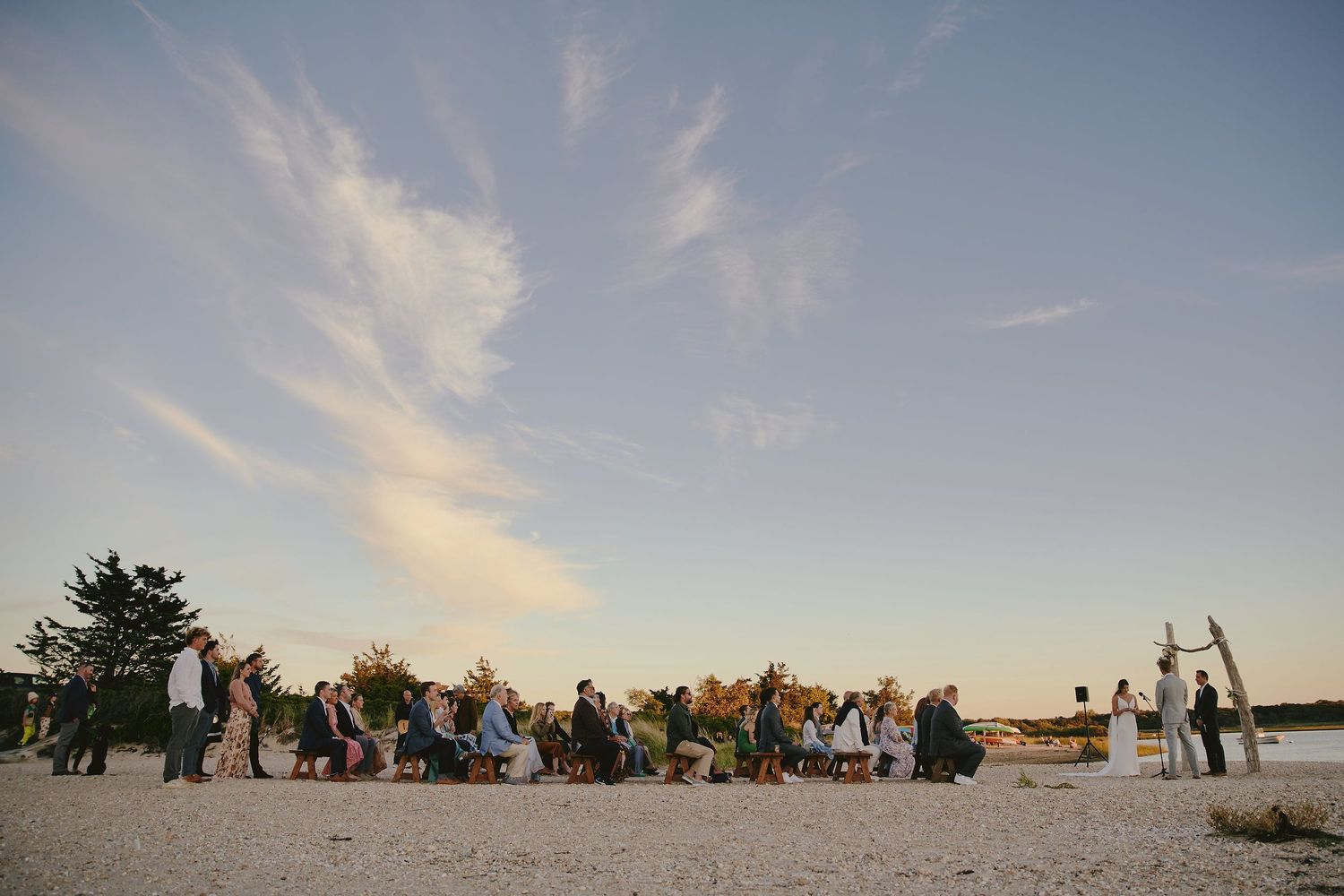Wedding guests gather on a sandy beach at sunset, creating a scenic outdoor celebration atmosphere.