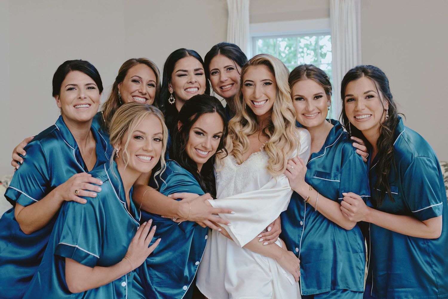 A bridal party in matching teal silk robes huddles together for a joyful pre-wedding photo.