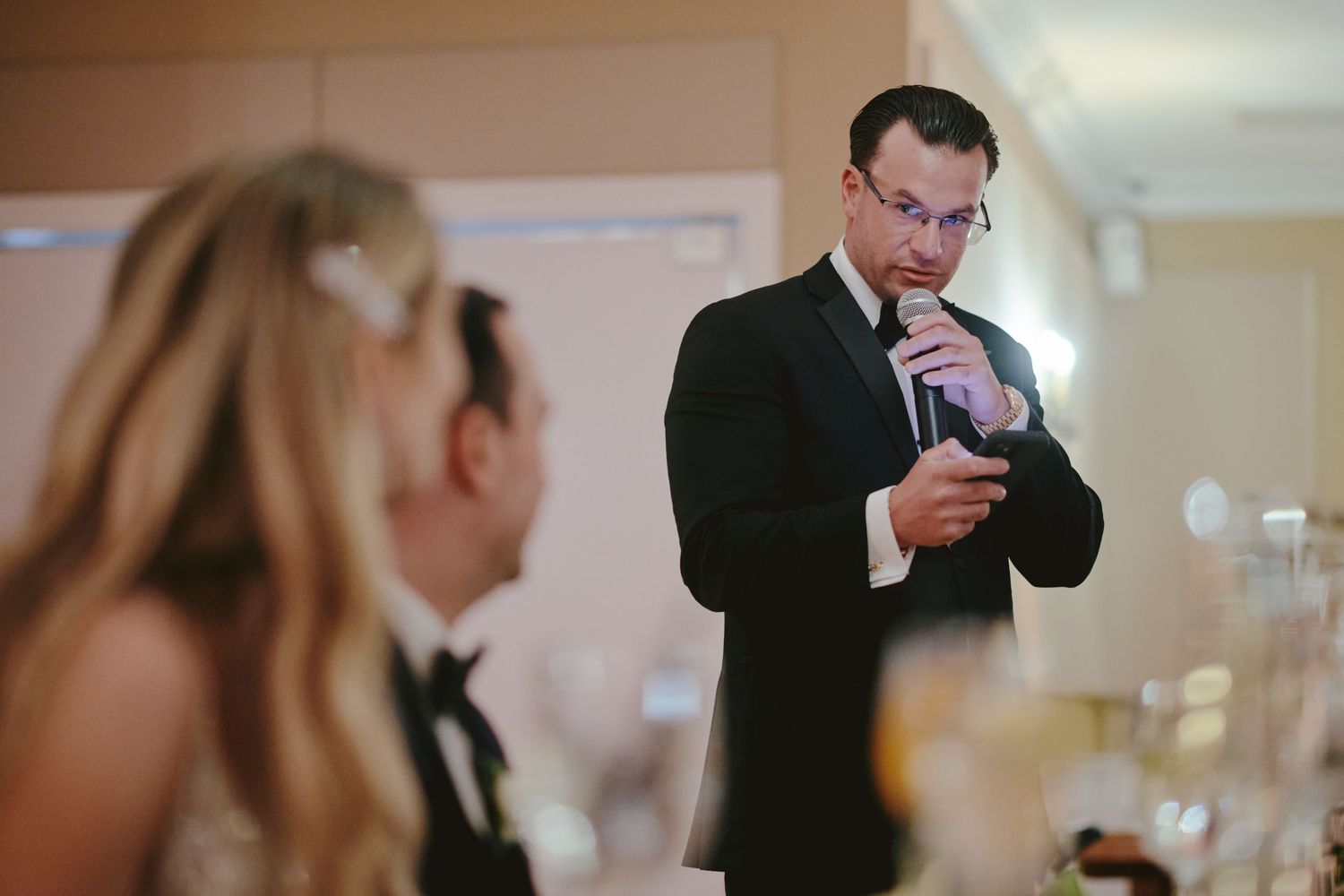 At a formal event, a person in a dark suit looks down intently at their phone while another attendee is nearby.