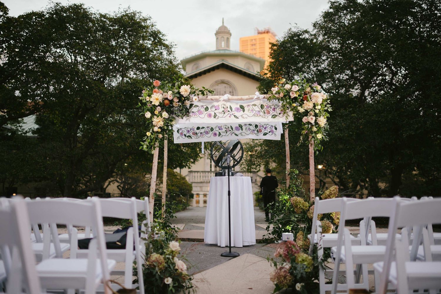 Outdoor wedding ceremony setup with floral-adorned arch, white chairs, and gazebo in background during golden hour.