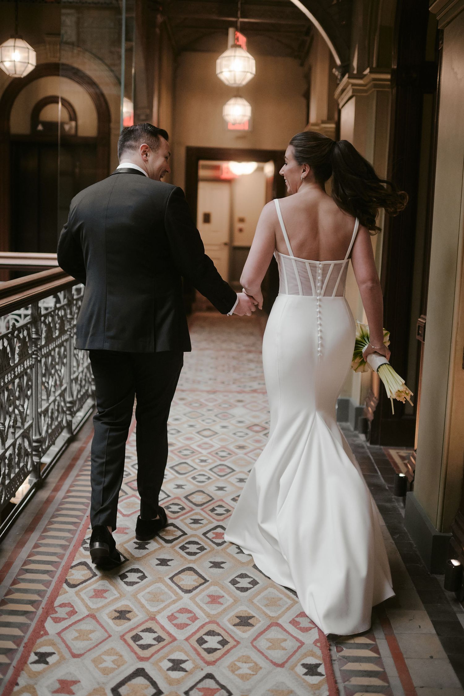 A couple walks hand in hand down an elegant hallway with ornate tiled flooring and hanging light fixtures.