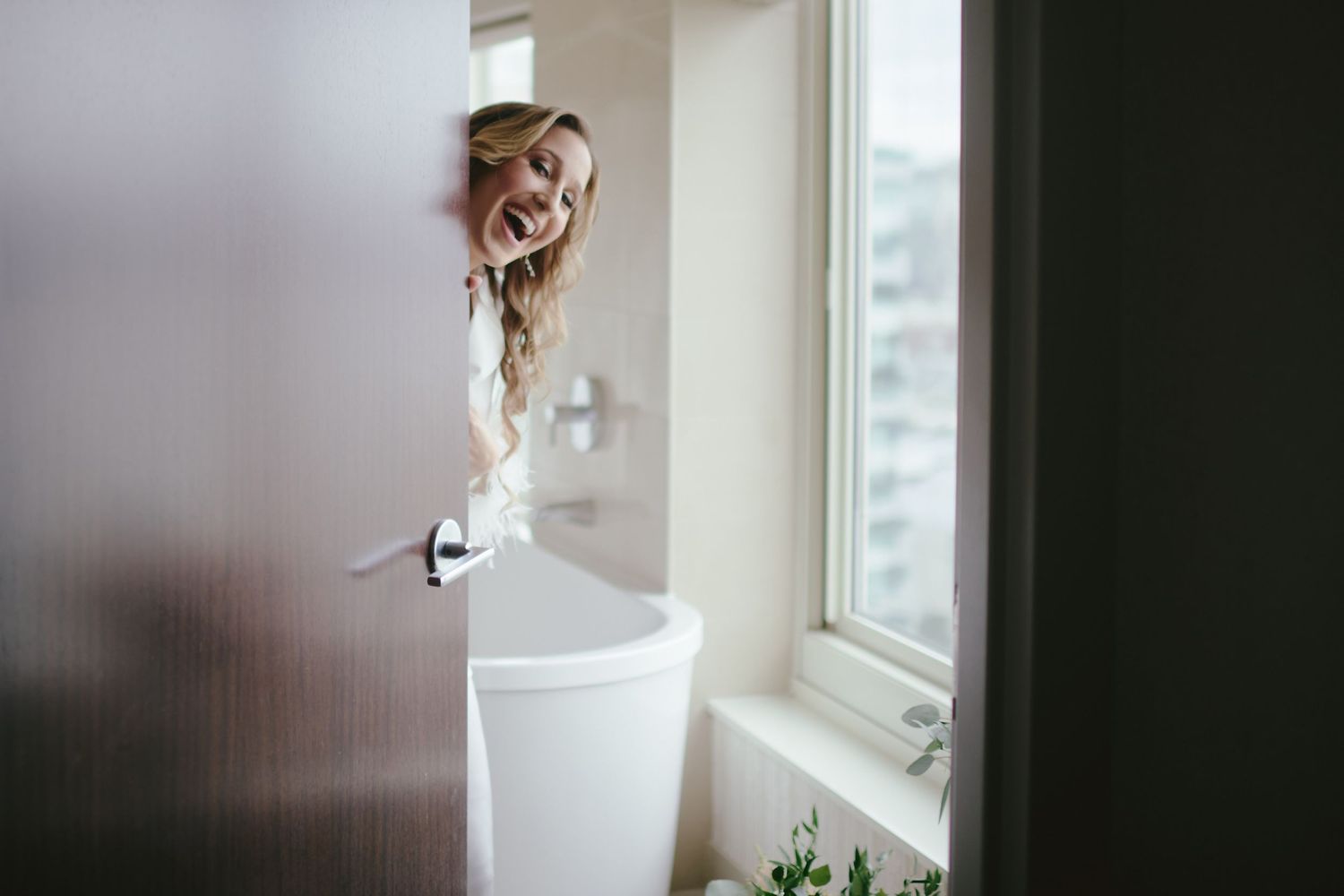 Person peeking playfully from behind a bathroom door near a white pedestal sink and window.