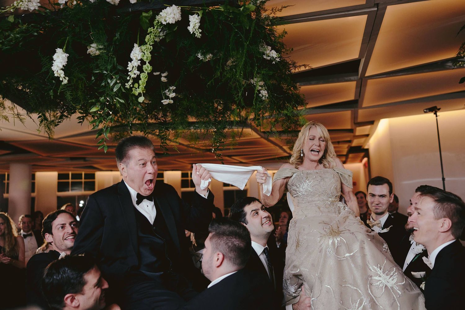Wedding celebration at an indoor reception venue with greenery and white floral decorations hanging from the ceiling.