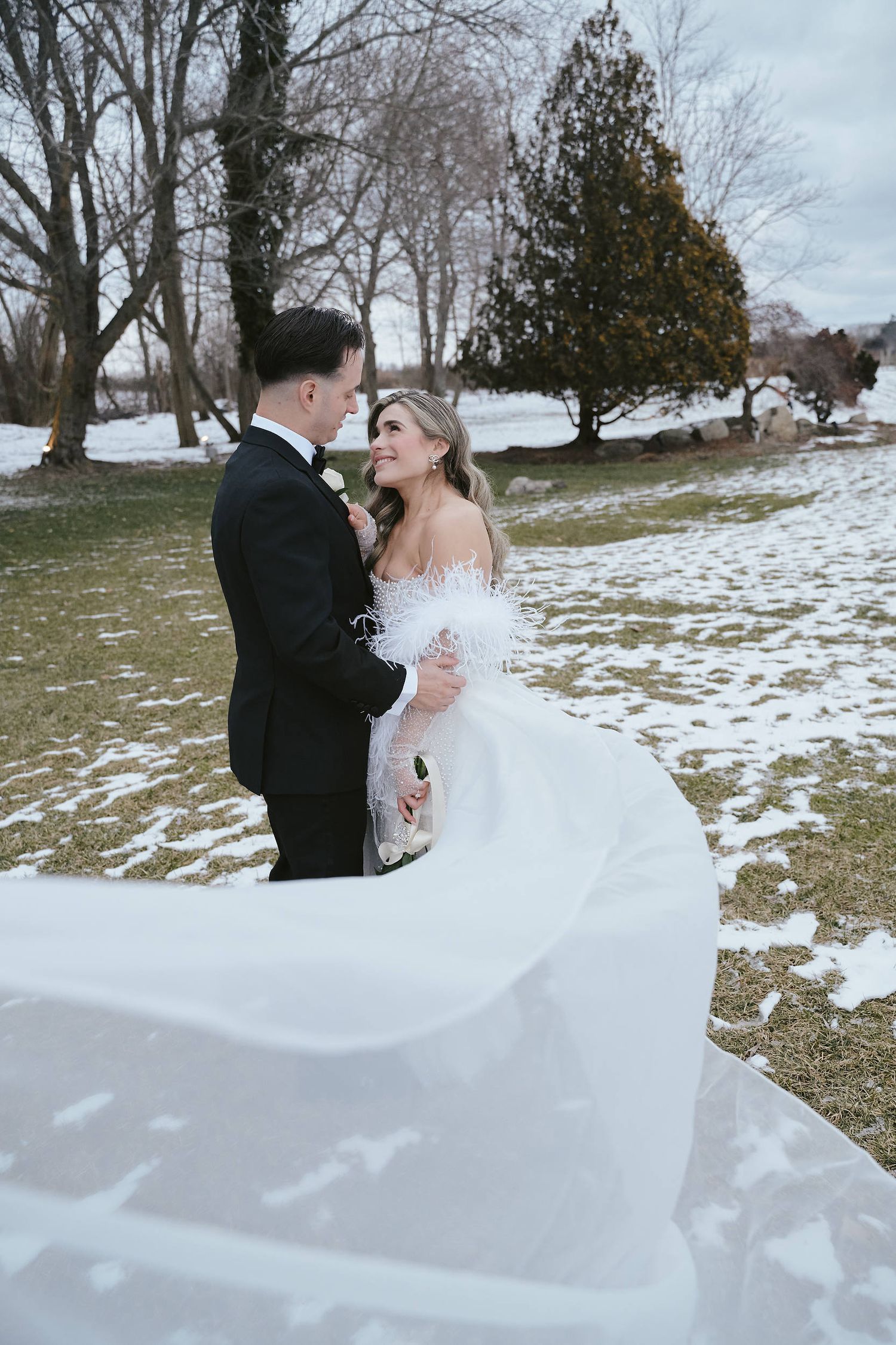 A winter wedding couple embraces in a snowy landscape with bare trees and patches of white ground in the background.