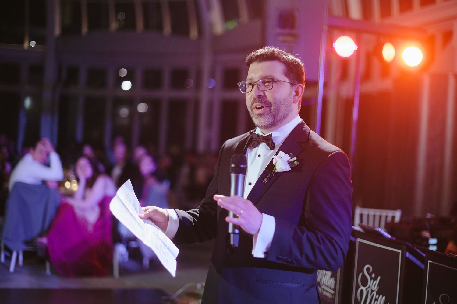 A speaker in formal attire delivers a speech at an evening event with stage lighting in the background.