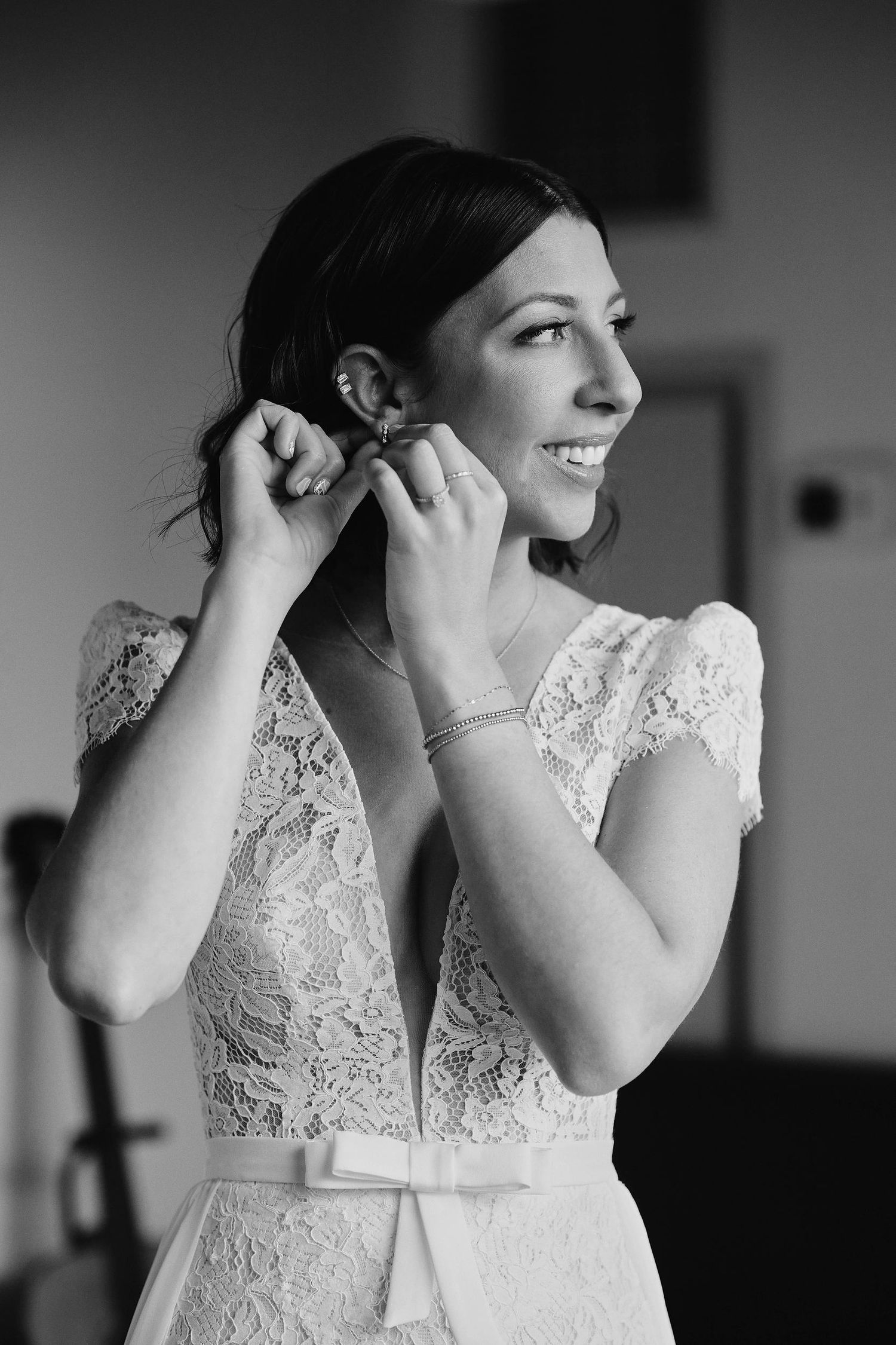 A bride in a lace wedding dress puts on her earrings while smiling in this black and white portrait.