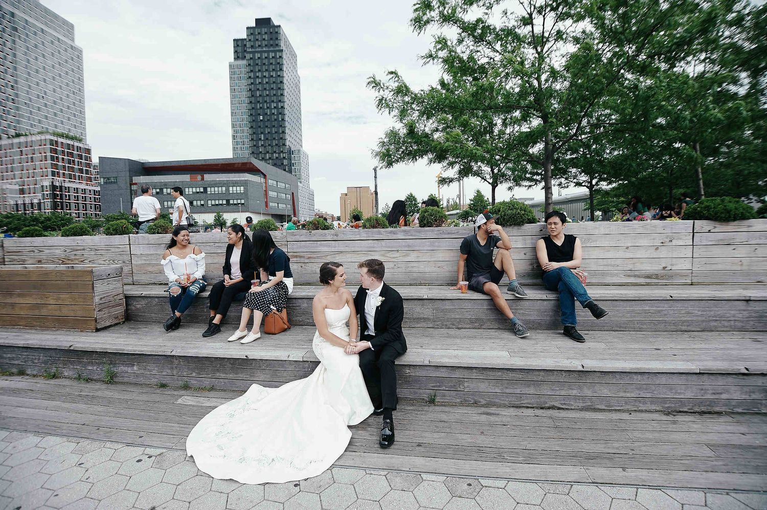 Couples relax on outdoor concrete steps at Long Island City waterfront with Manhattan skyline and trees in background.