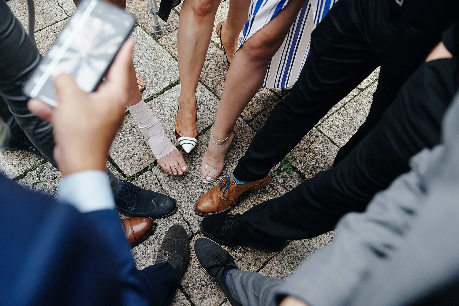 Wedding guests place their feet in a circle formation for a creative group photo on stone pavement.
