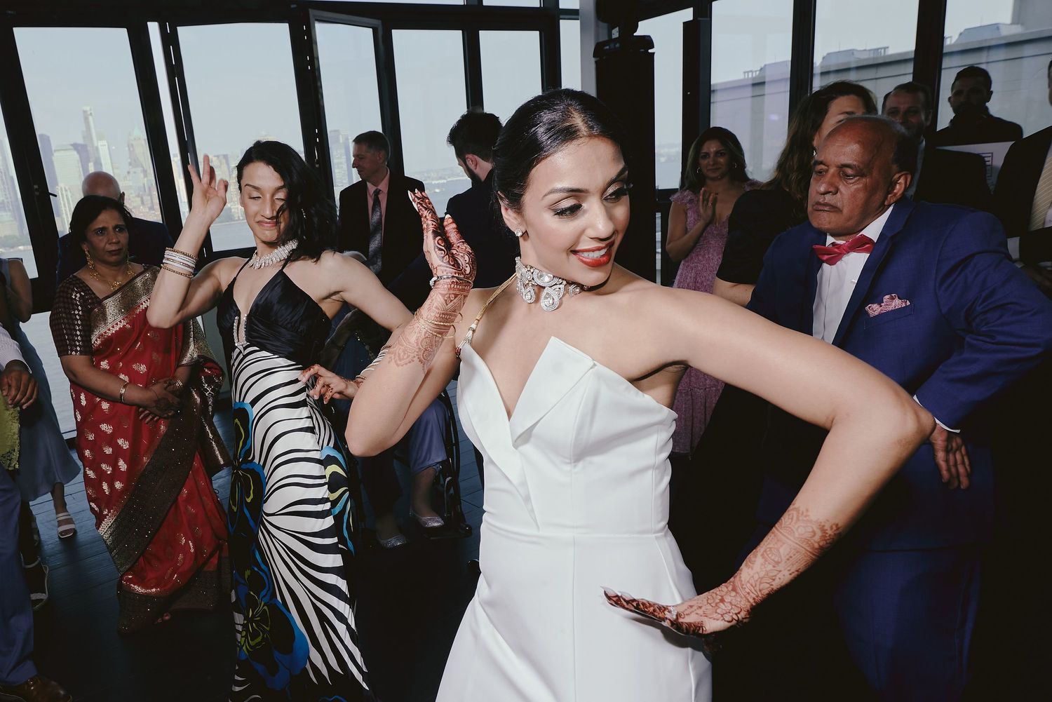 Wedding guests dancing and celebrating at a reception in front of large windows.