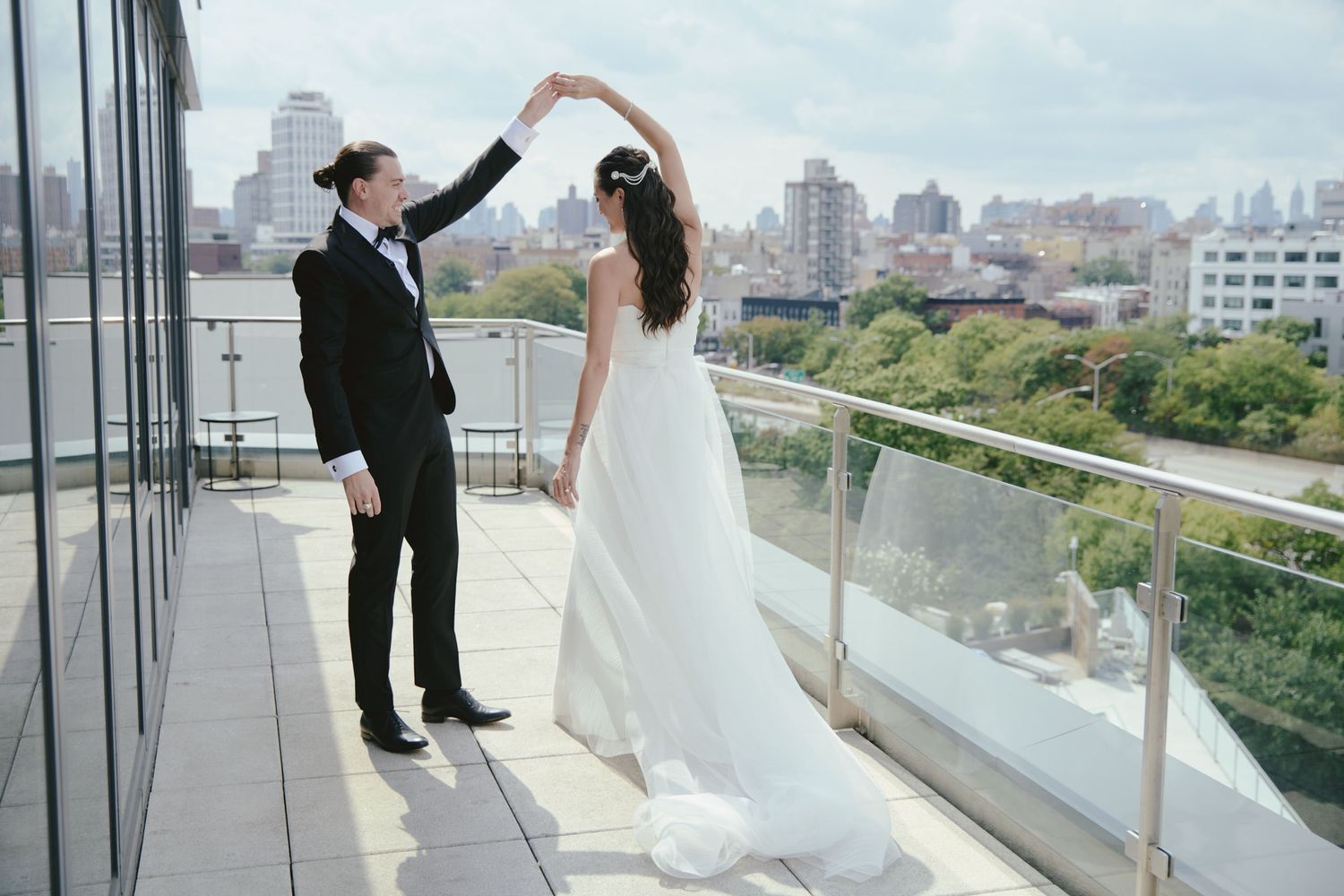 Two people in wedding attire dancing together on a balcony overlooking a city skyline on a sunny day.
